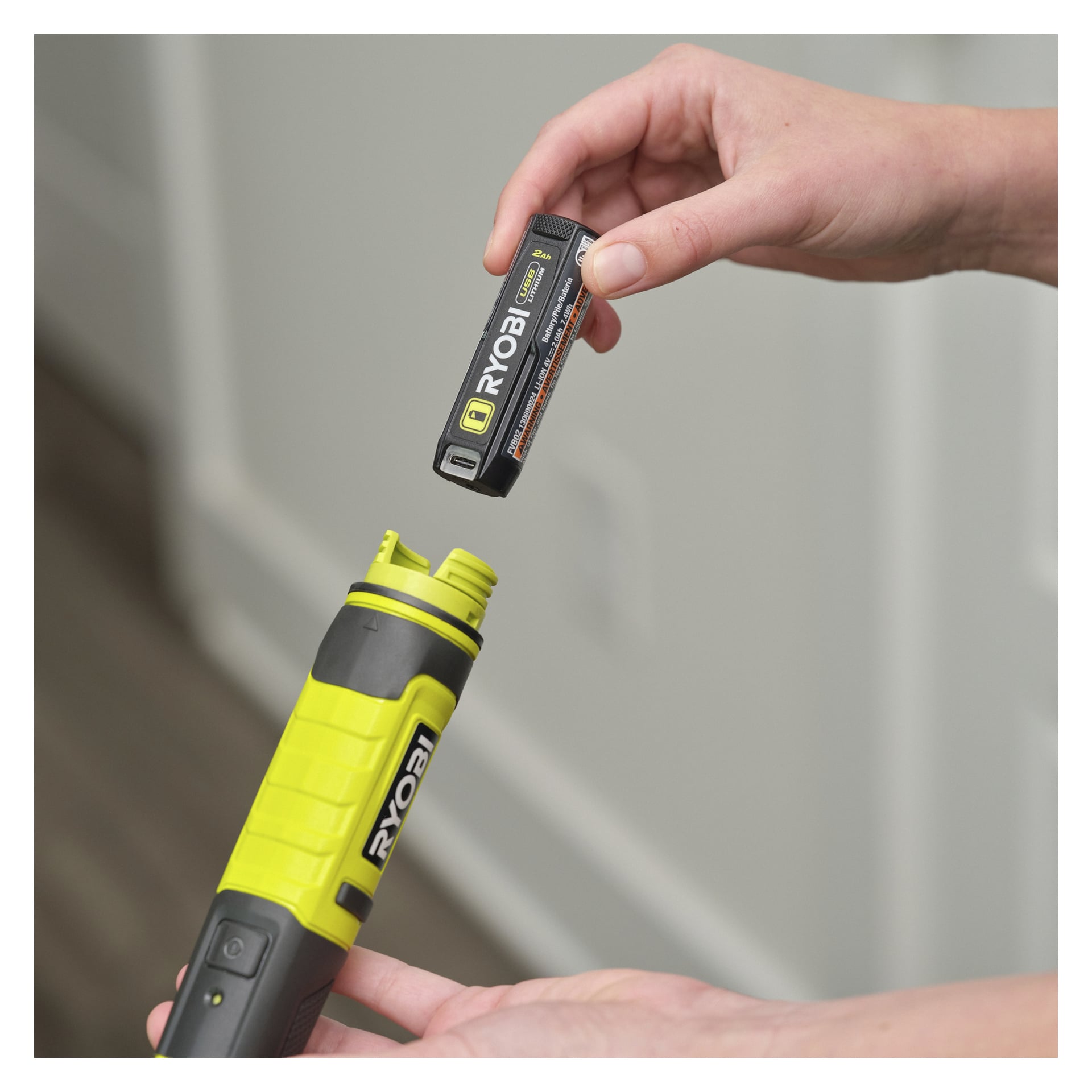 En person placerar ett Ryobi USB Lithium-batteri i handtaget på en gul och svart RDS4-0 detaljrengöringsborste inomhus.