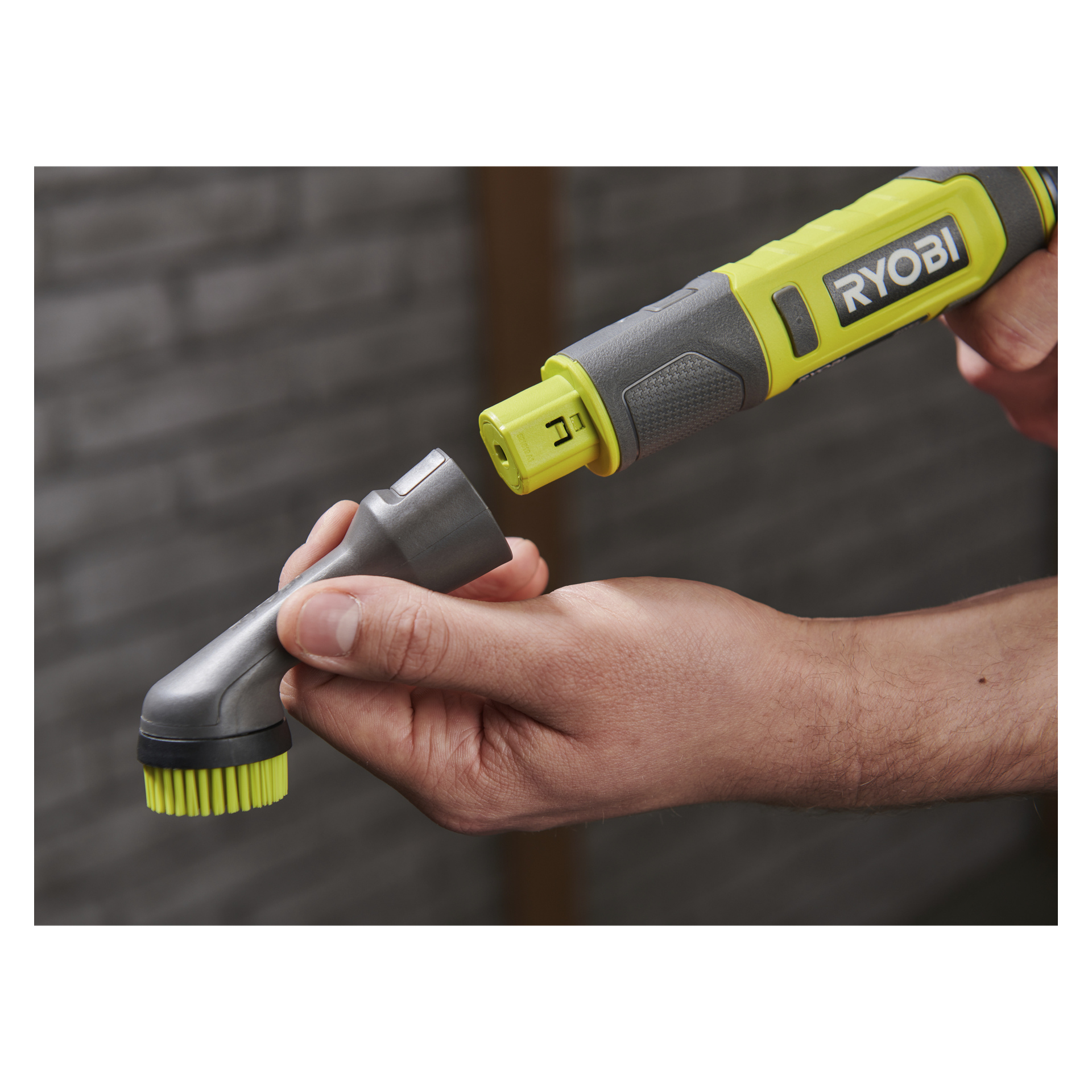 Någon kopplar ett borsthuvud till en Ryobi USB litiumbatteri detaljrengöringsborste, med en tegelvägg mjukt suddig bakom dem.