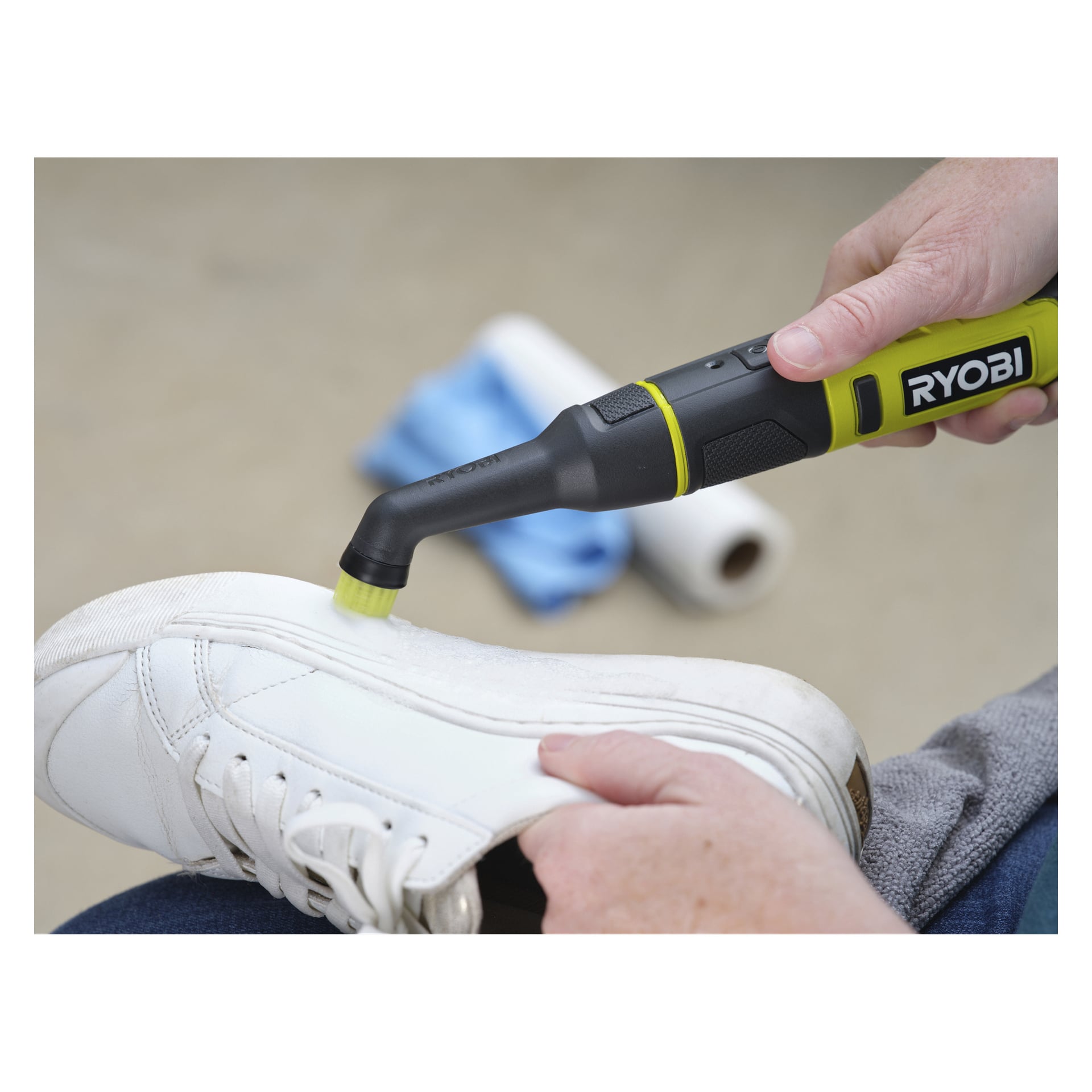 En person rengör en vit sneaker med en Ryobi USB Lithium detaljborste; En pappersrulle och en blå trasa finns i bakgrunden.