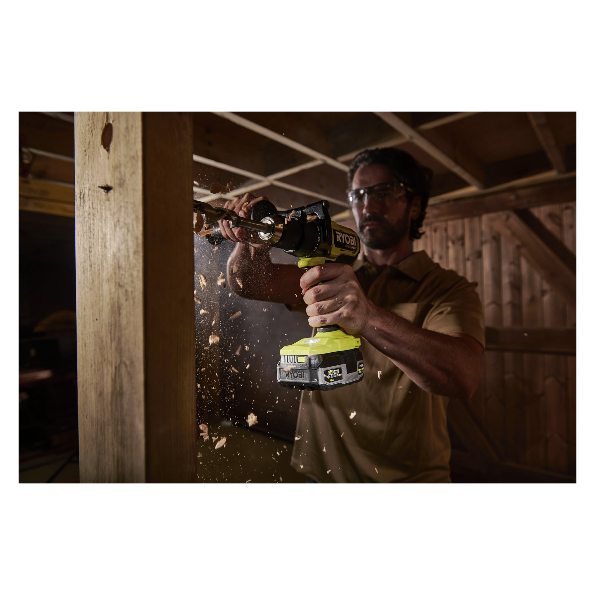 En person med skyddsglasögon borrar i en träbalk med en Ryobi One+ HP Battery Edge-RB1840E och sprider träspån.