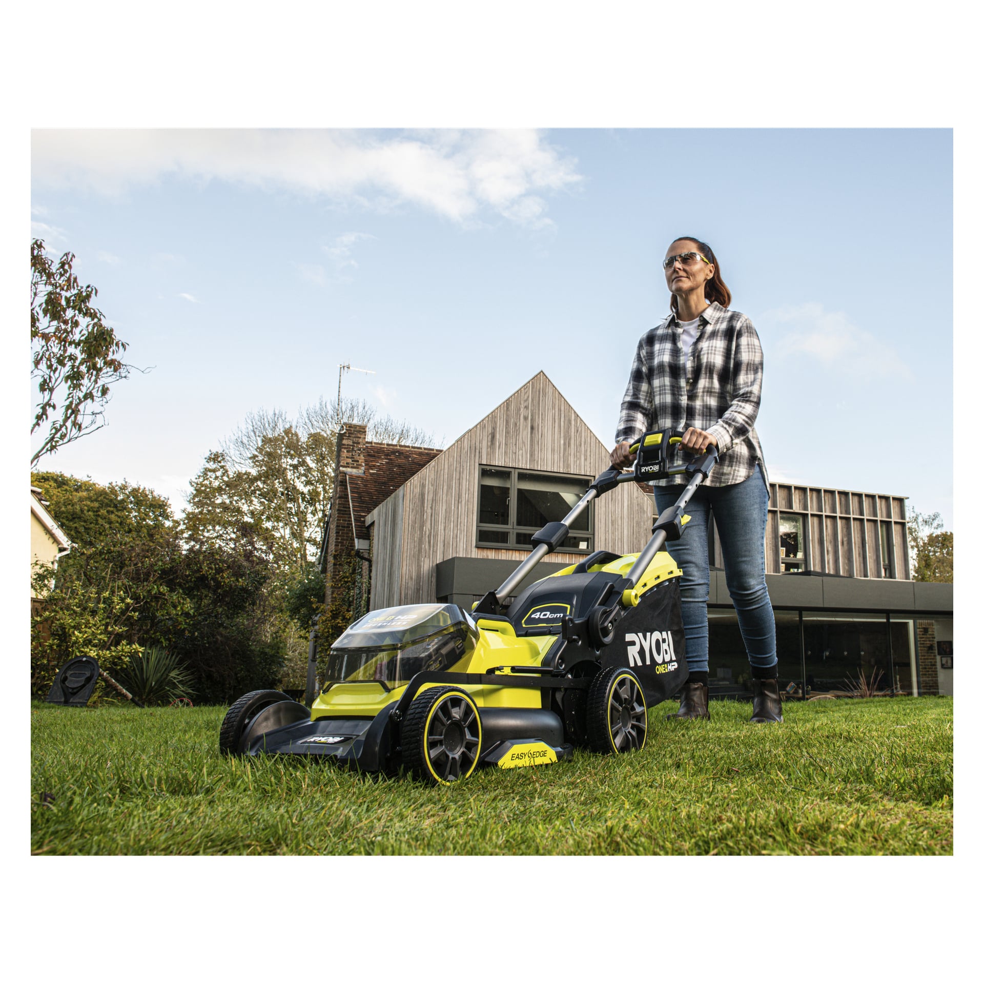 Med solglasögon och rutig skjorta klipper en person en gräsbevuxen gräsmatta med en Ryobi One+ HP Battery Edge-RB1840E nära ett modernt hus.