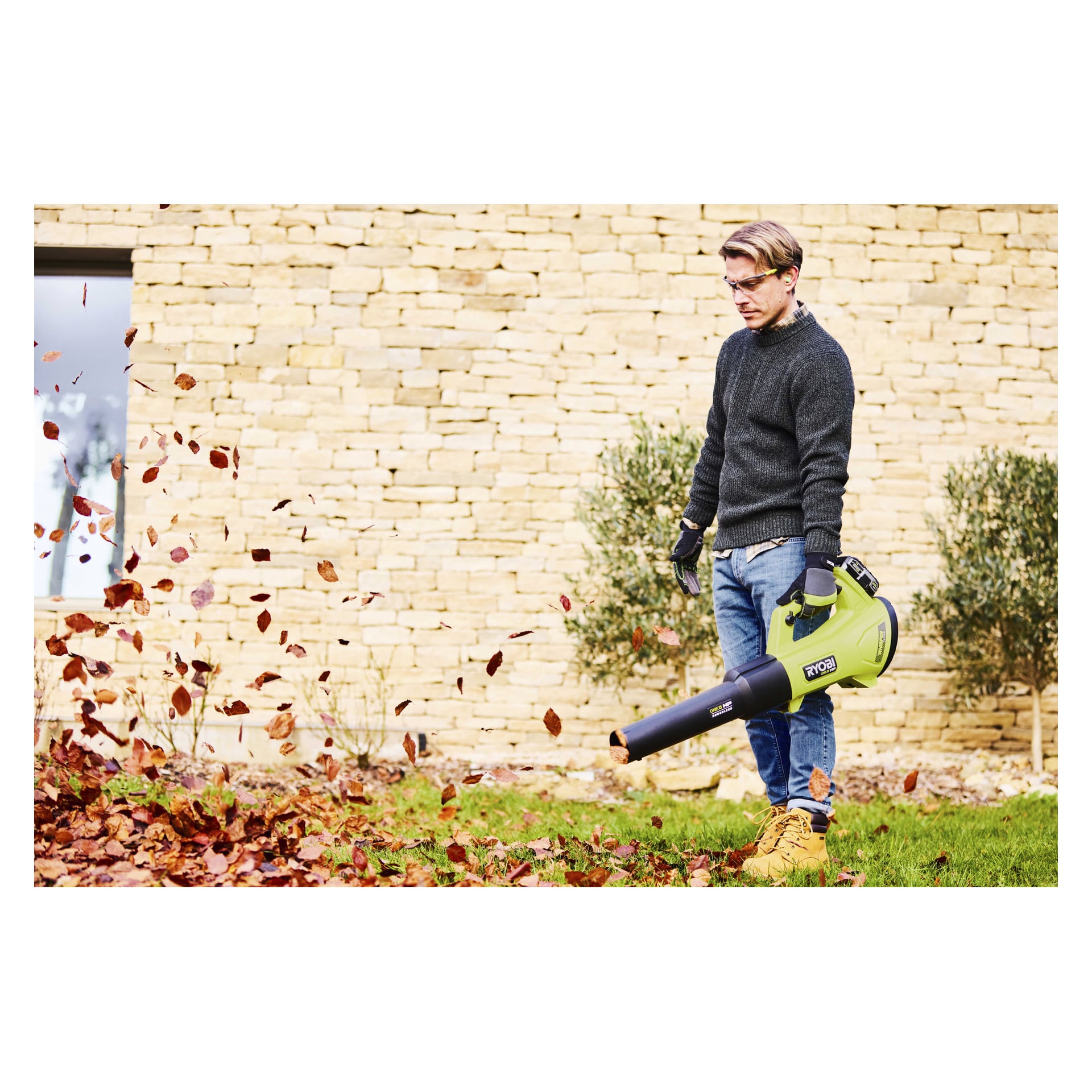 Någon som bär en mörk tröja, jeans, handskar och glasögon använder en Ryobi One+ HP lövblåsare på gräs nära en stenmur och växt.