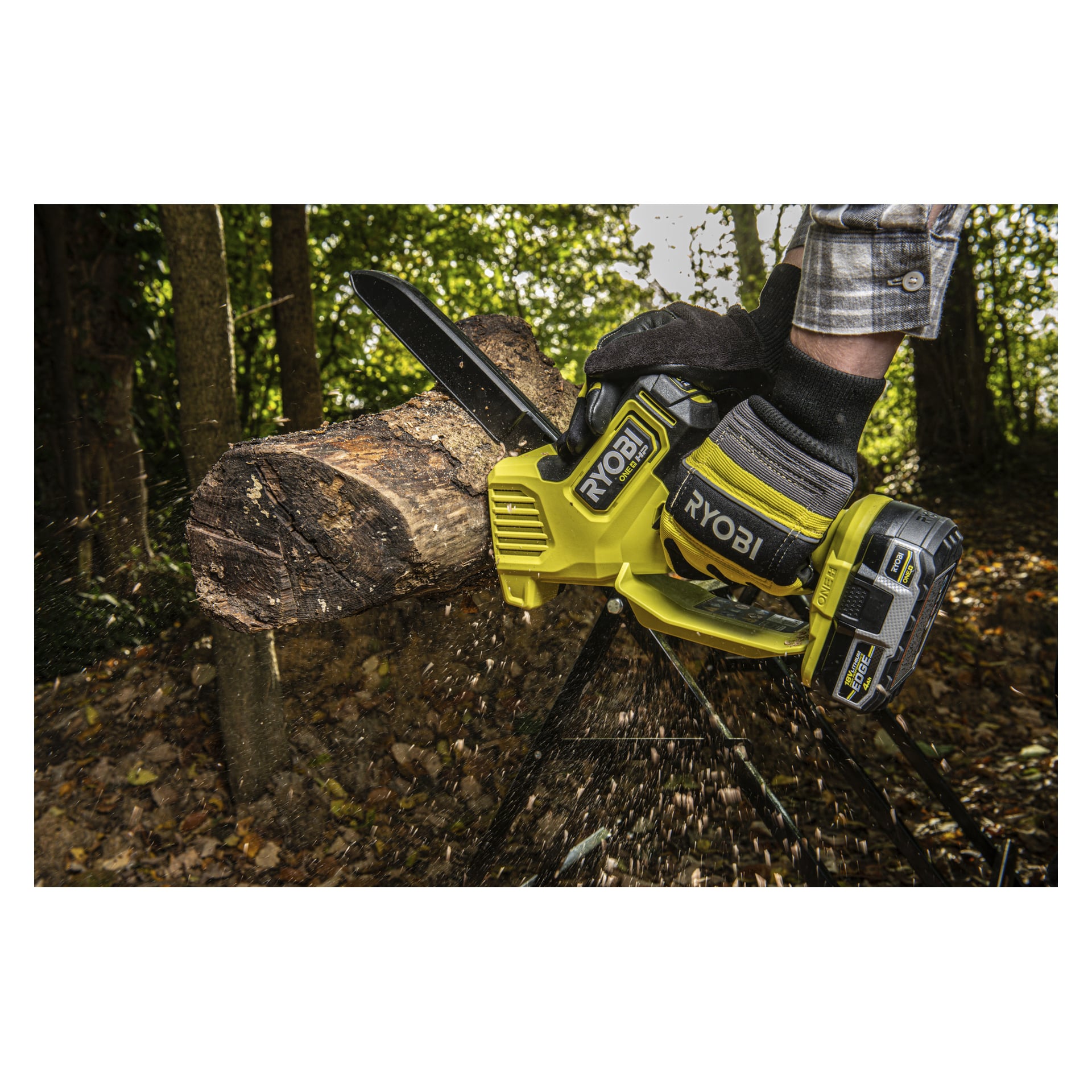 En person i svarta handskar hugger en stock på en sågbock utanför med en gul Ryobi One+ HP motorsåg, sågspån flyger och träd bakom.
