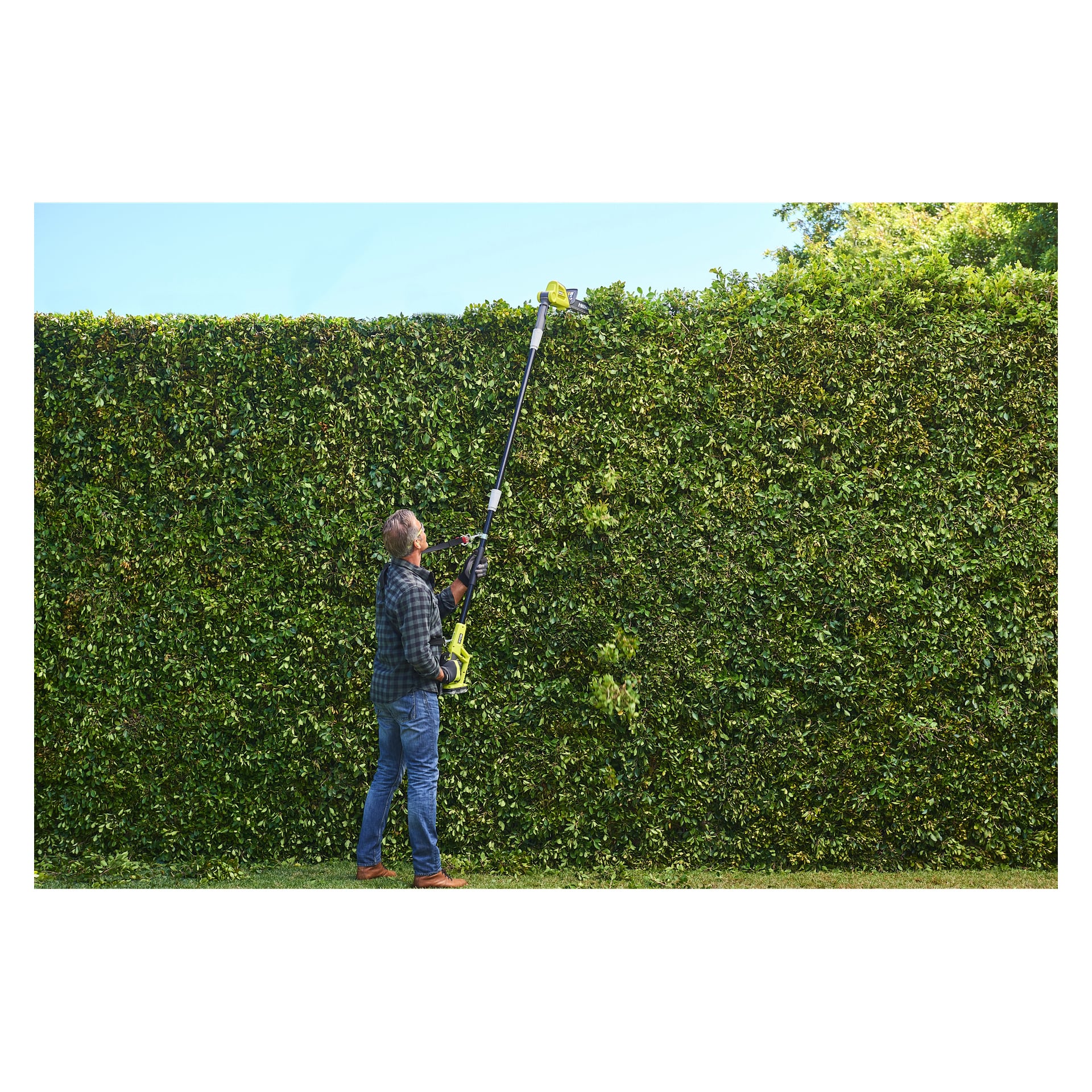 En person på gräs under en klar himmel klipper den övre delen av en hög, tjock grön häck med en batteridriven Ryobi One+ häcksax.