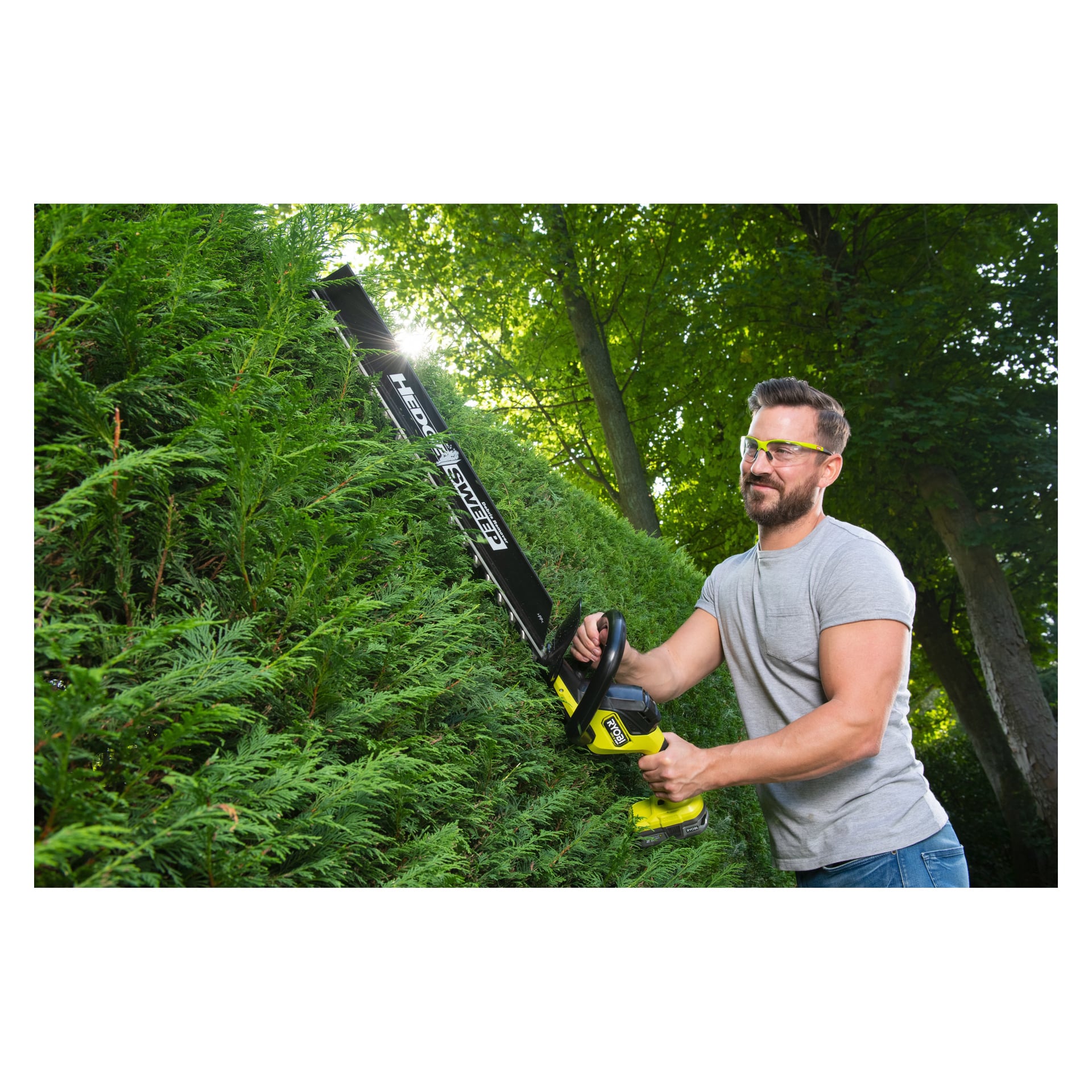 En man med skyddsglasögon och grå t-shirt trimmar en häck utomhus med en Ryobi RY18HTX60A-220 trimmer på en solig, grön dag.
