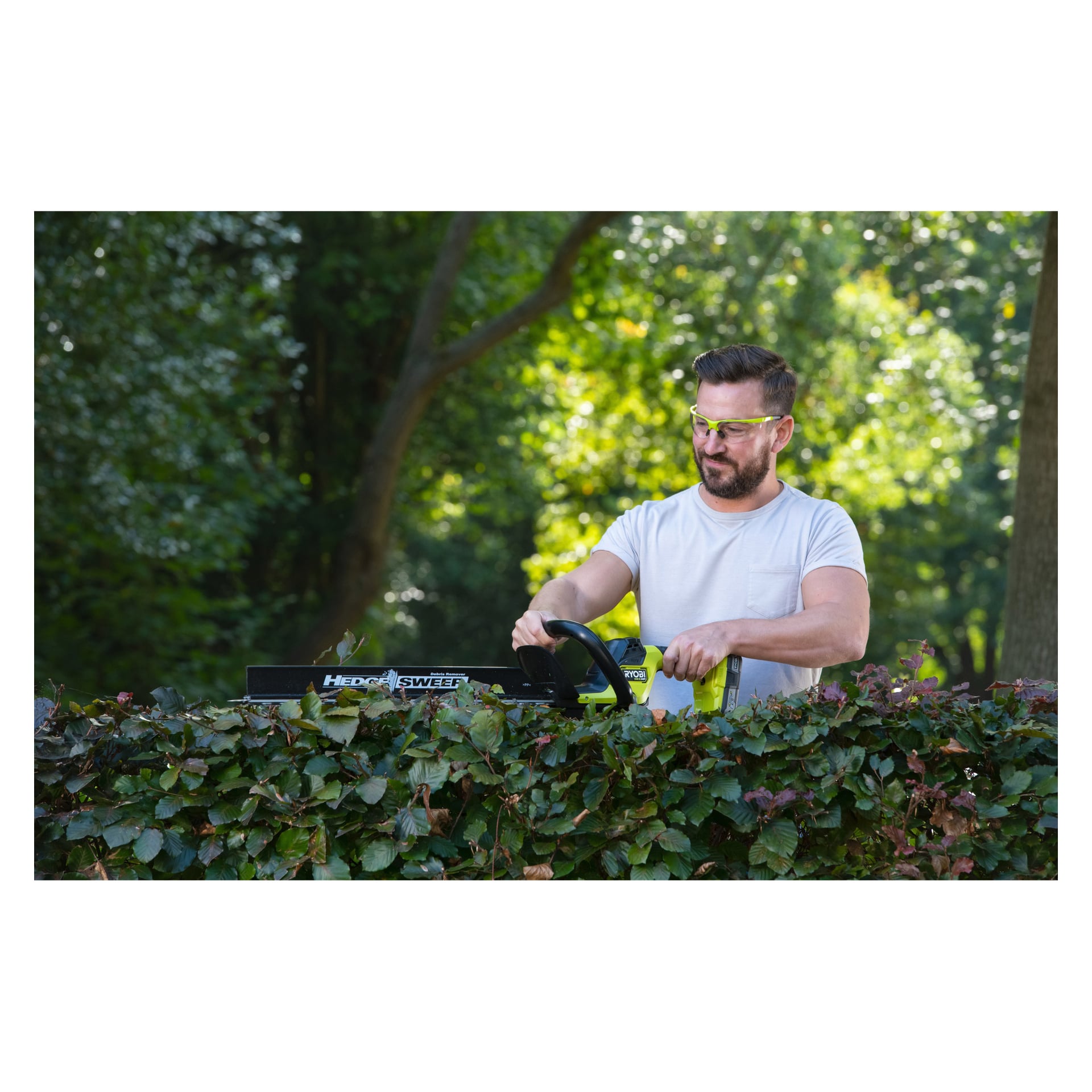 En man med skyddsglasögon och en vit t-shirt använder en Ryobi RY18HTX60A-220 batteridriven häcksax för att forma buskar bland gröna träd.