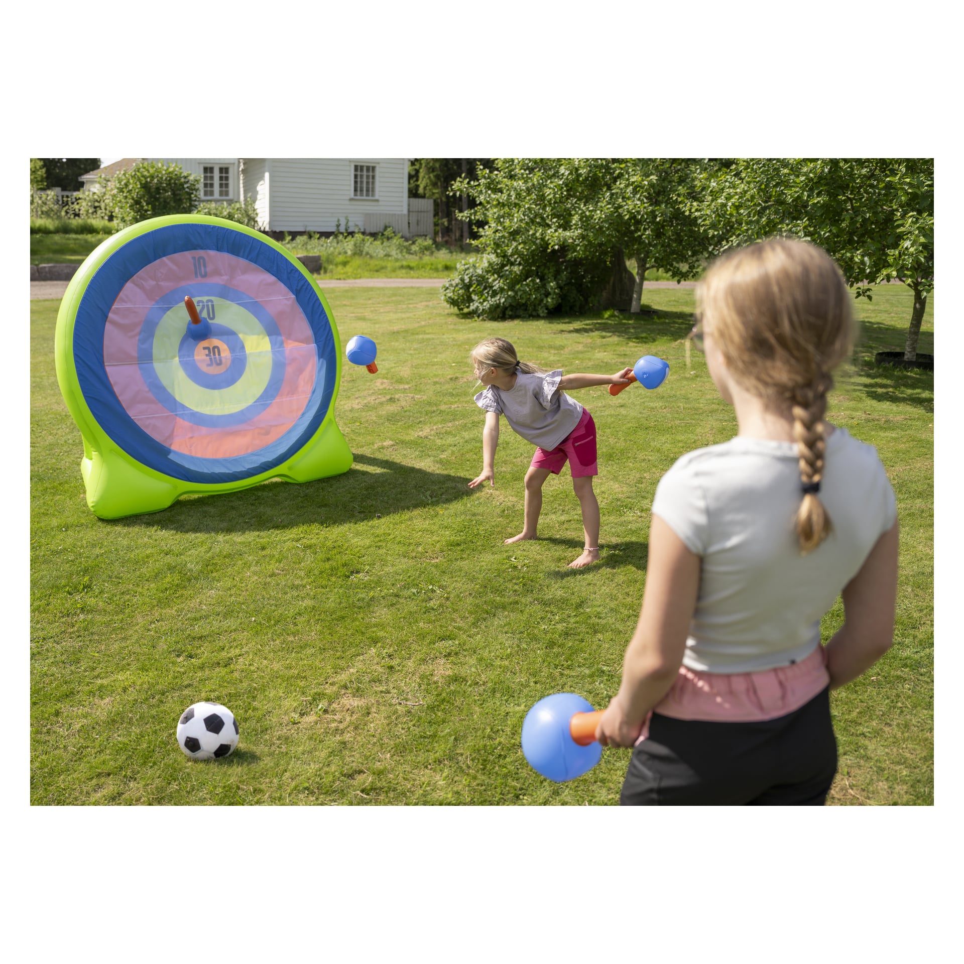 Kaksi lasta pelaa ulkona Bestway Inflatable Dartboard Backyard Bullseye 2-in-1 -peliä, tähtäämässä pehmeillä nuolia isoon ilmatäytteiseen kohteeseen nurmikolla. Toinen lapsi on kesken heiton, kun taas toinen seisoo pitelemässä tikkaa. Jalkapallo on nurmikolla, ja taustalla näkyy puita ja talo.