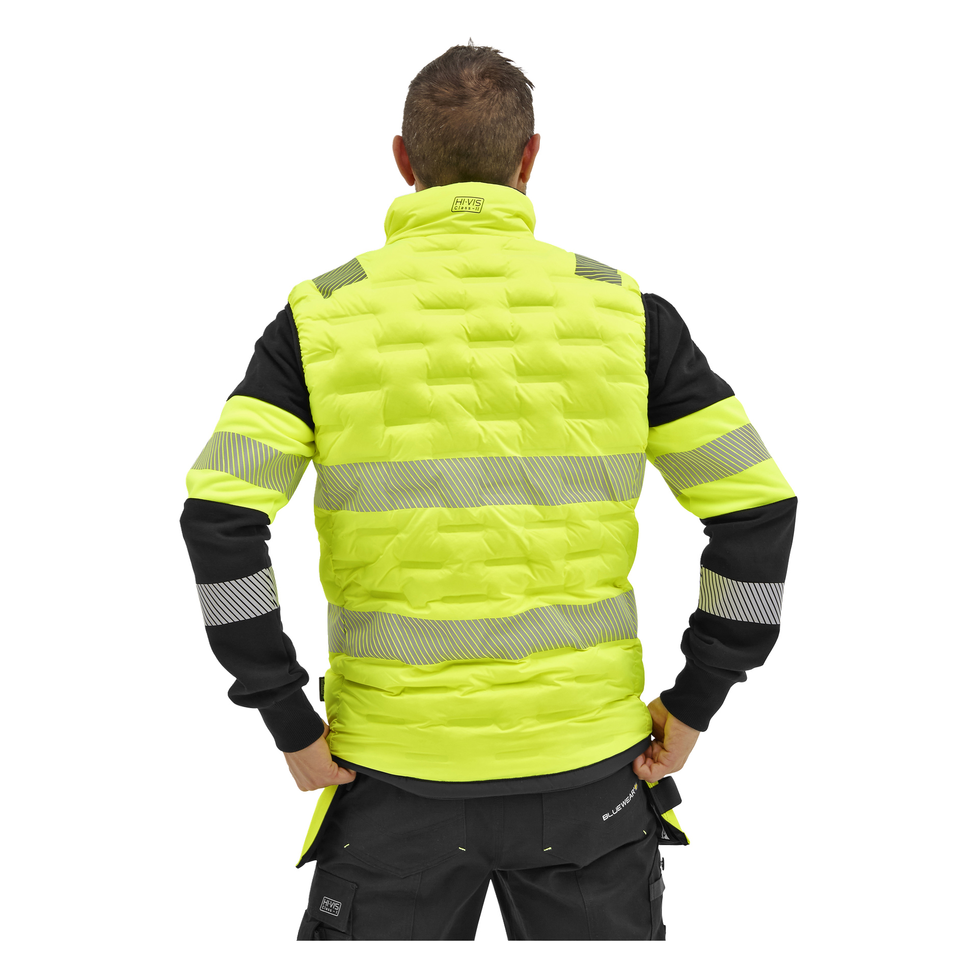En person vänder sig bort, händerna på höfterna, och bär en Bluewear Warm-fodrad vändbar hög-vis-väst från Edmonton med reflekterande ränder över svarta kläder.