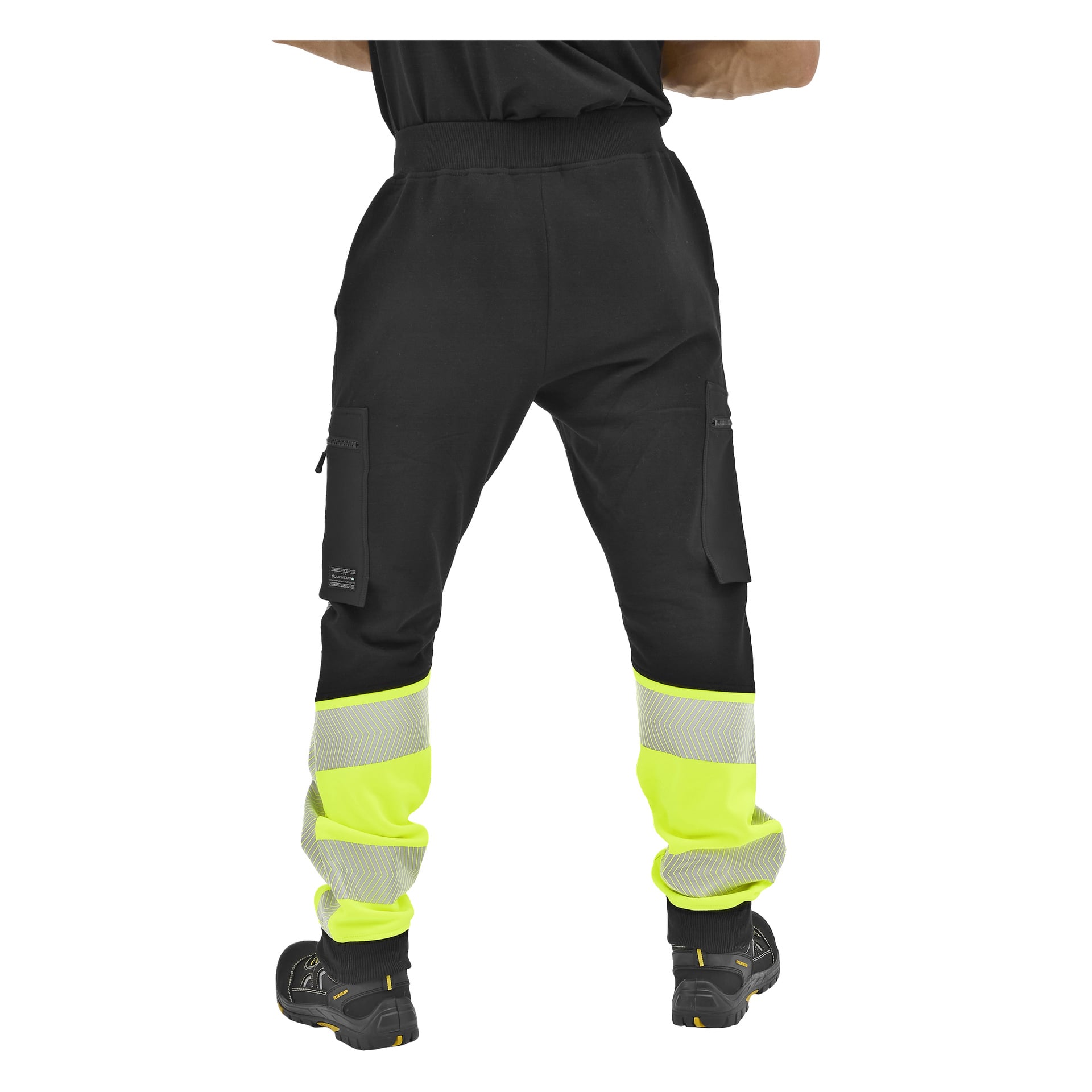 En person står med ryggen mot kameran, bär svarta Bluewear högsynliga byxor med gula reflekterande band och svarta skor.