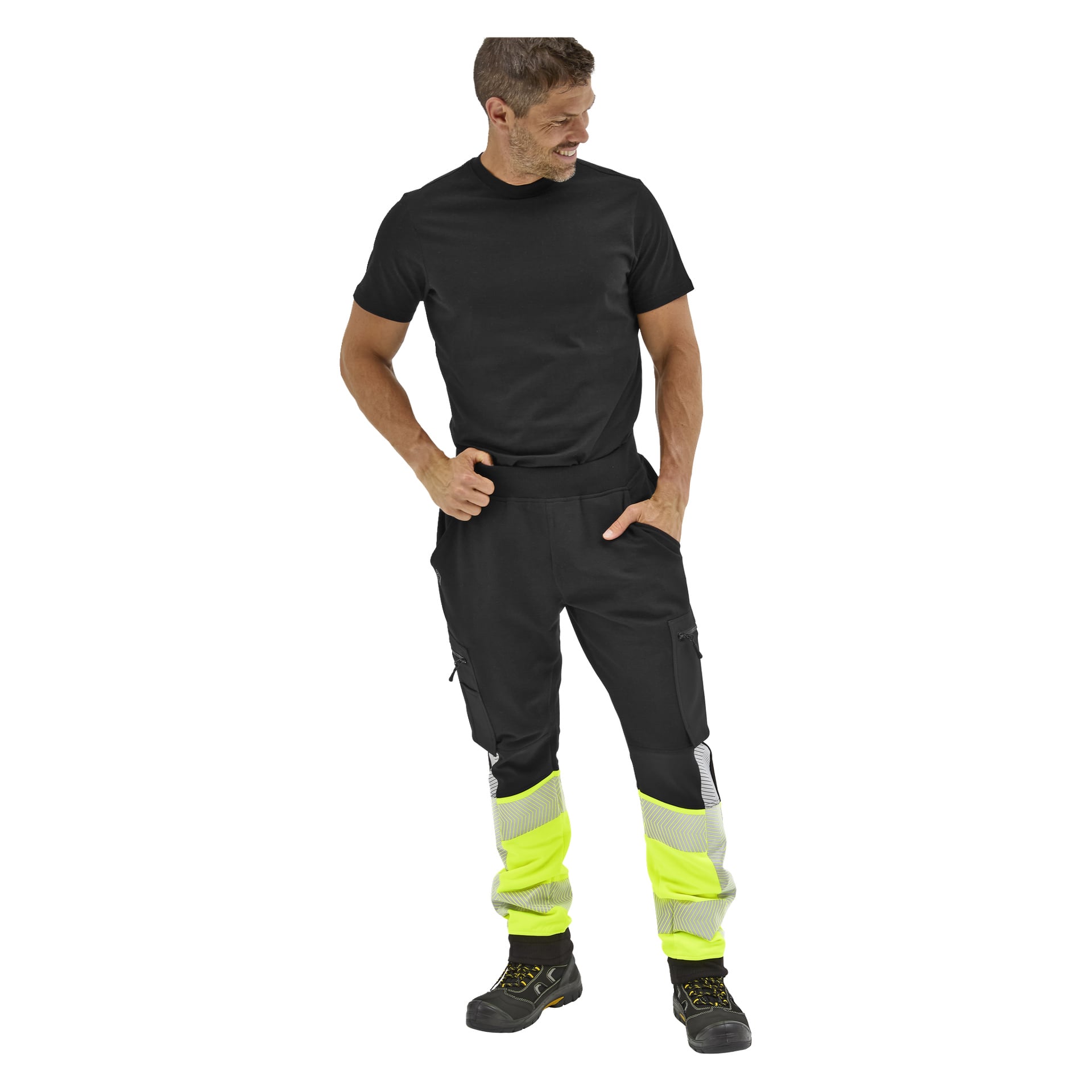 En man i Bluewear Essex högvarbyxor, svart t-shirt och svarta arbetsskor med gula detaljer står leende och kastar en blick åt sidan.