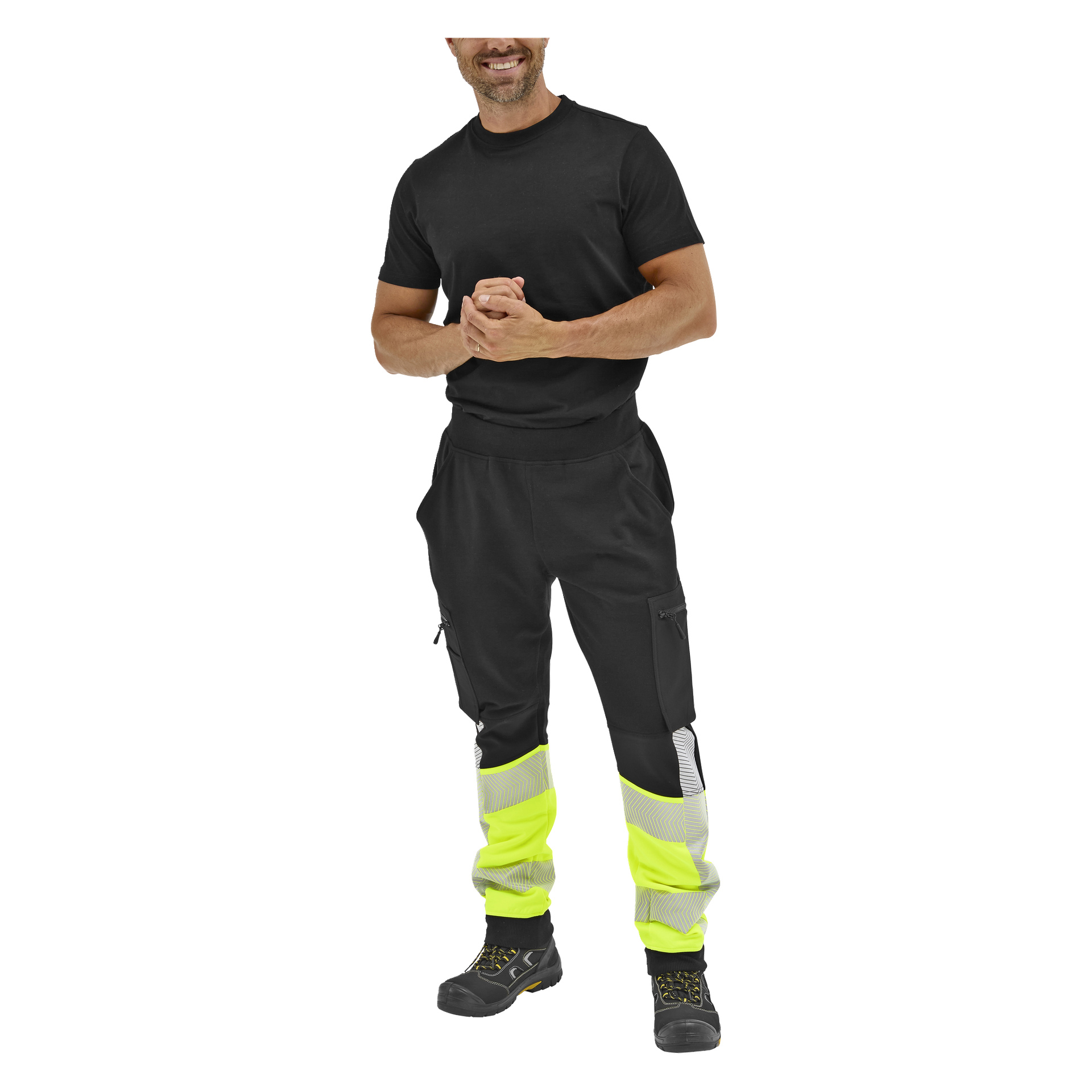 En leende man står med händerna knäppta, klädd i en svart t-shirt, neongula byxor med reflexband och svarta stövlar.
