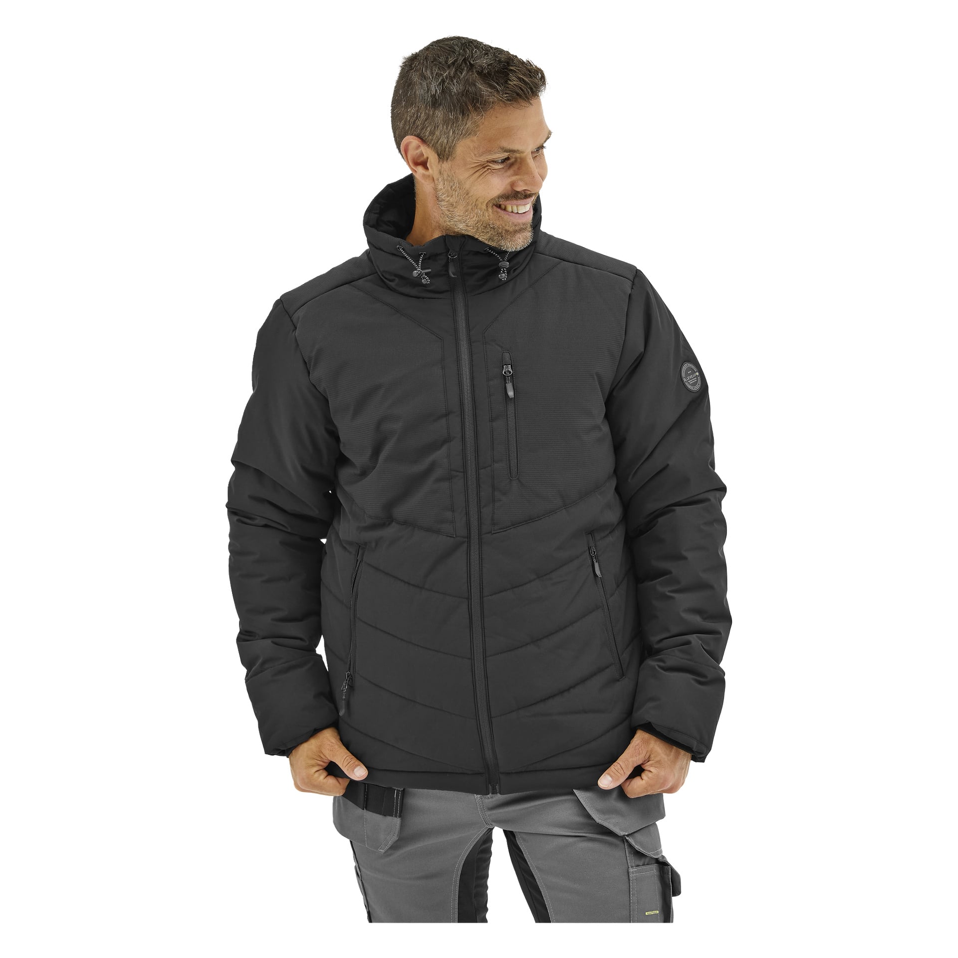 En man ler i en svart Bluewear Folke-vadderad jacka med huva och dragkedjefickor, tillsammans med ljusgrå byxor med svarta detaljer.
