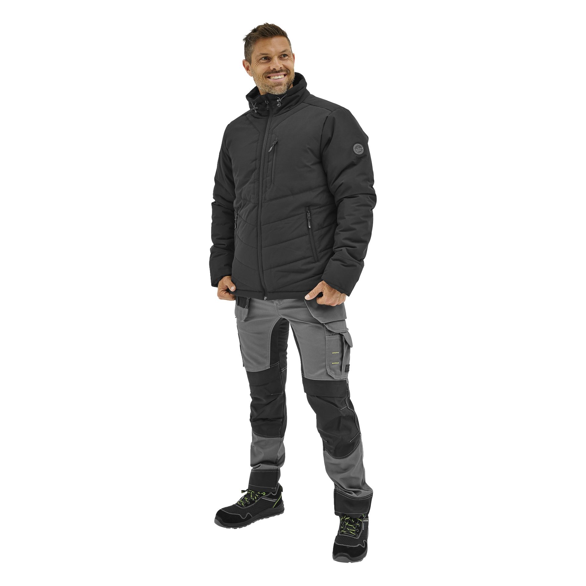 En leende man bär en Bluewear Padded Men's Jacket Folke, grå och svarta arbetsbyxor med fickor och svarta stövlar med gröna snören.