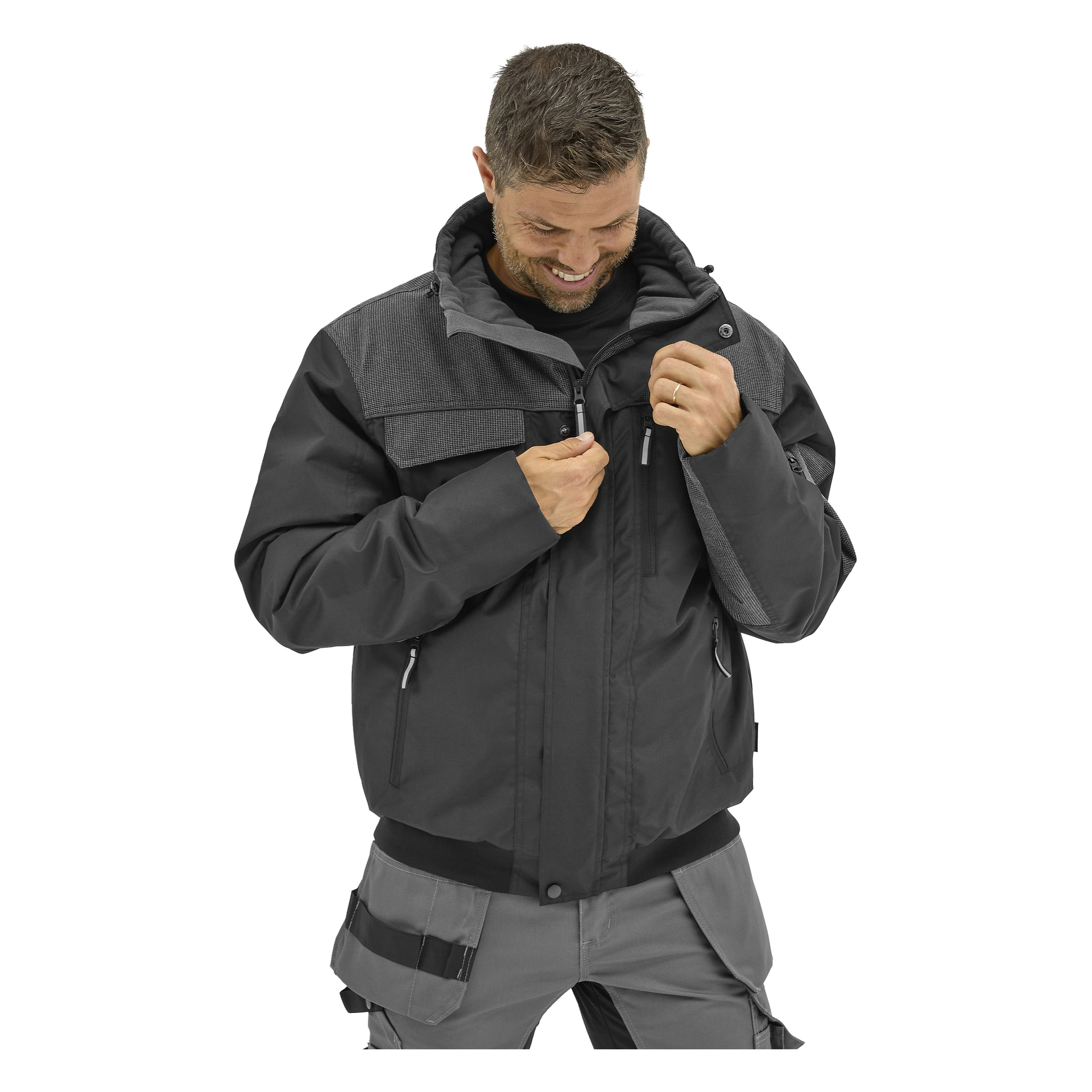 En man med kort hår drar igen dragkedjan på en svart och grå Bluewear Men's Johan arbetsjacka med flera fickor, tillsammans med grå arbetsbyxor.