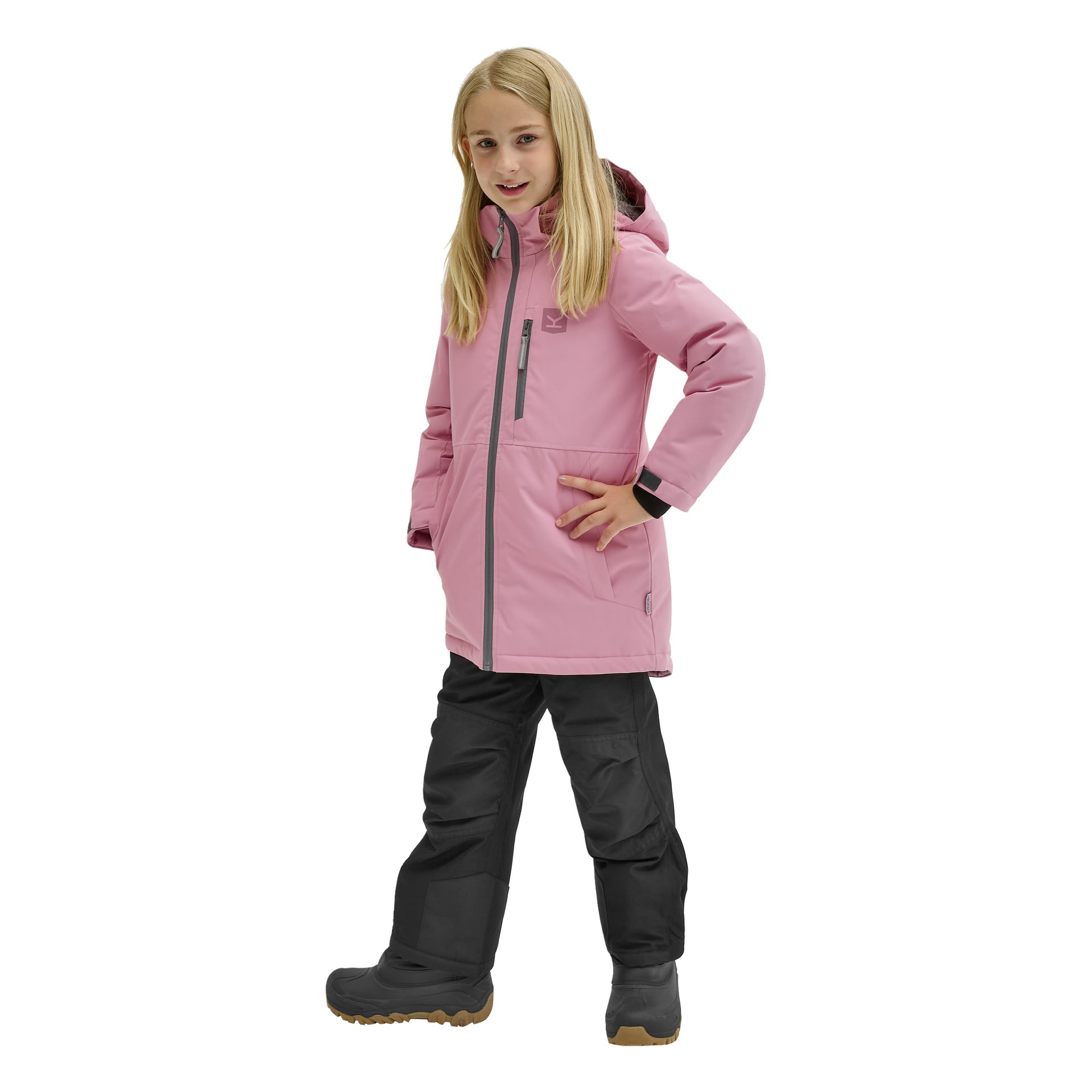 Ett barn med långt blont hår bär Kayoba Outdoor Children's vinterjacka Kläppen i rosa, tillsammans med svarta snöbyxor och bruna stövlar, och står självsäkert med ena handen på höften och den andra avslappnad vid sidan.