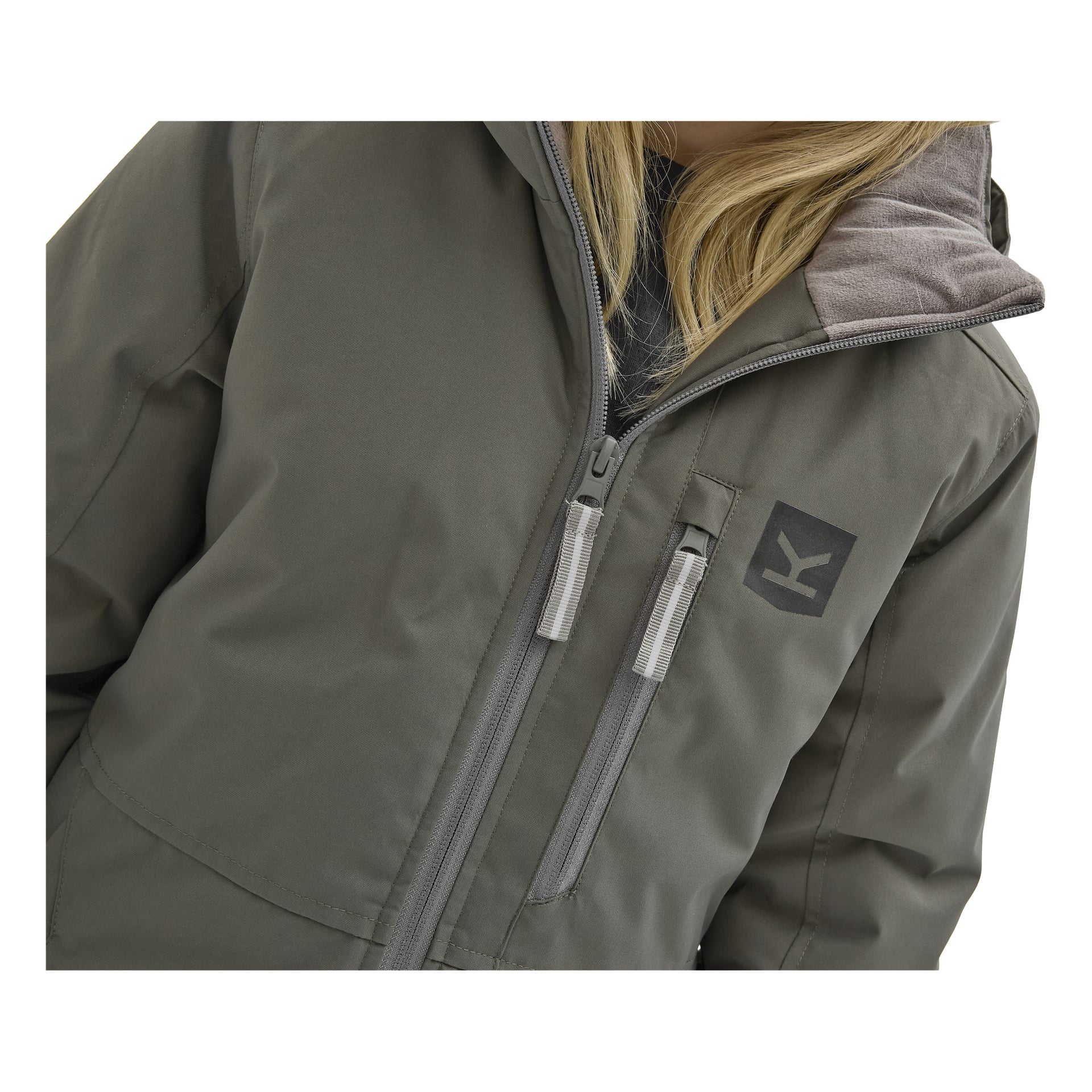 Närbild på en person som bär Kayoba Outdoor Children's vinterjacka Kläppen i grått med huva, dragkedja och en bröstlogotyp där det står "K." Personens blonda hår syns, men ansiktet syns inte.