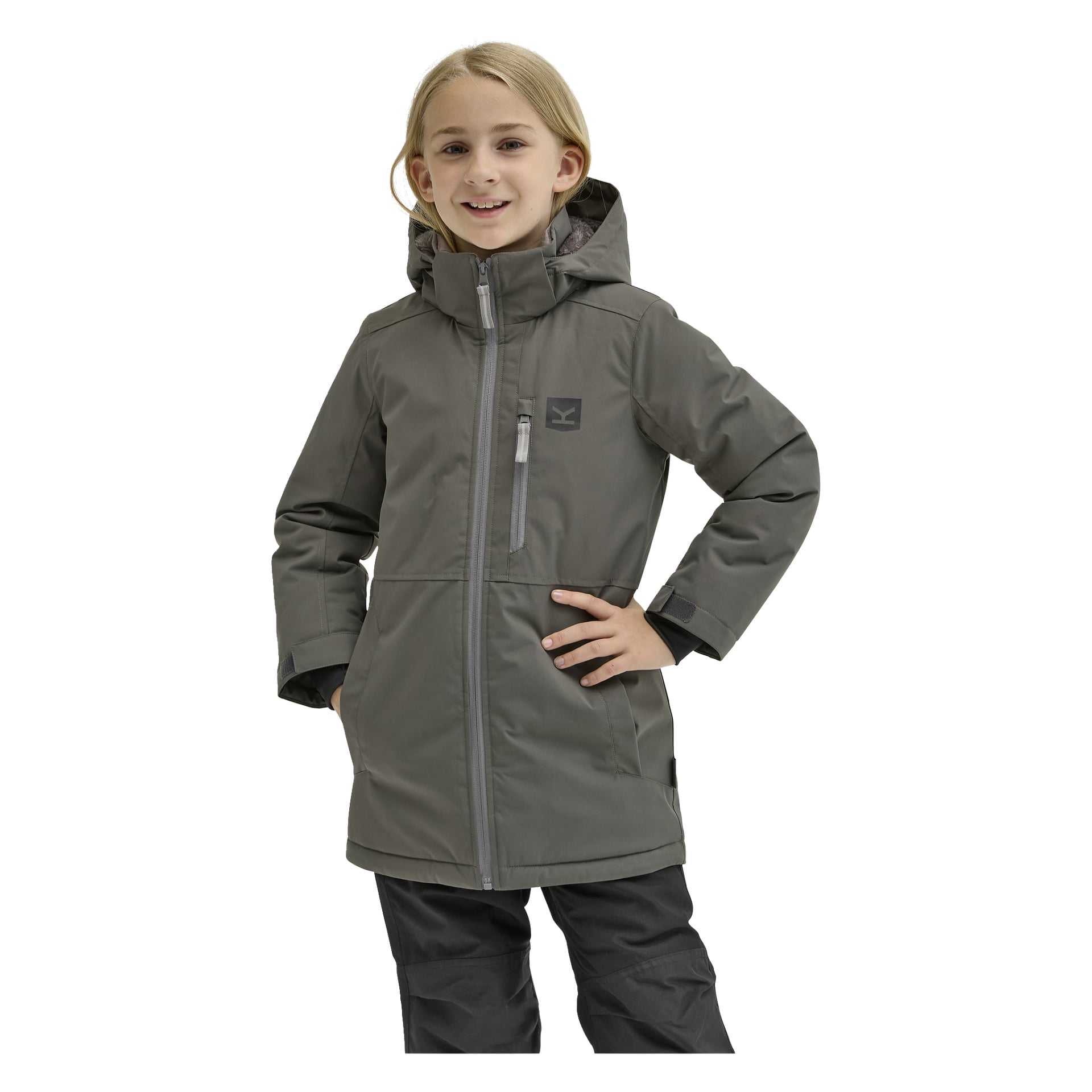 Ett leende barn med långt blont hår står med ena handen på höften, klädd i "Kayoba Outdoor Children's winter jacket Kläppen", en grå huva med dragkedja på bröstet och dragkedja fram, samt svarta byxor.