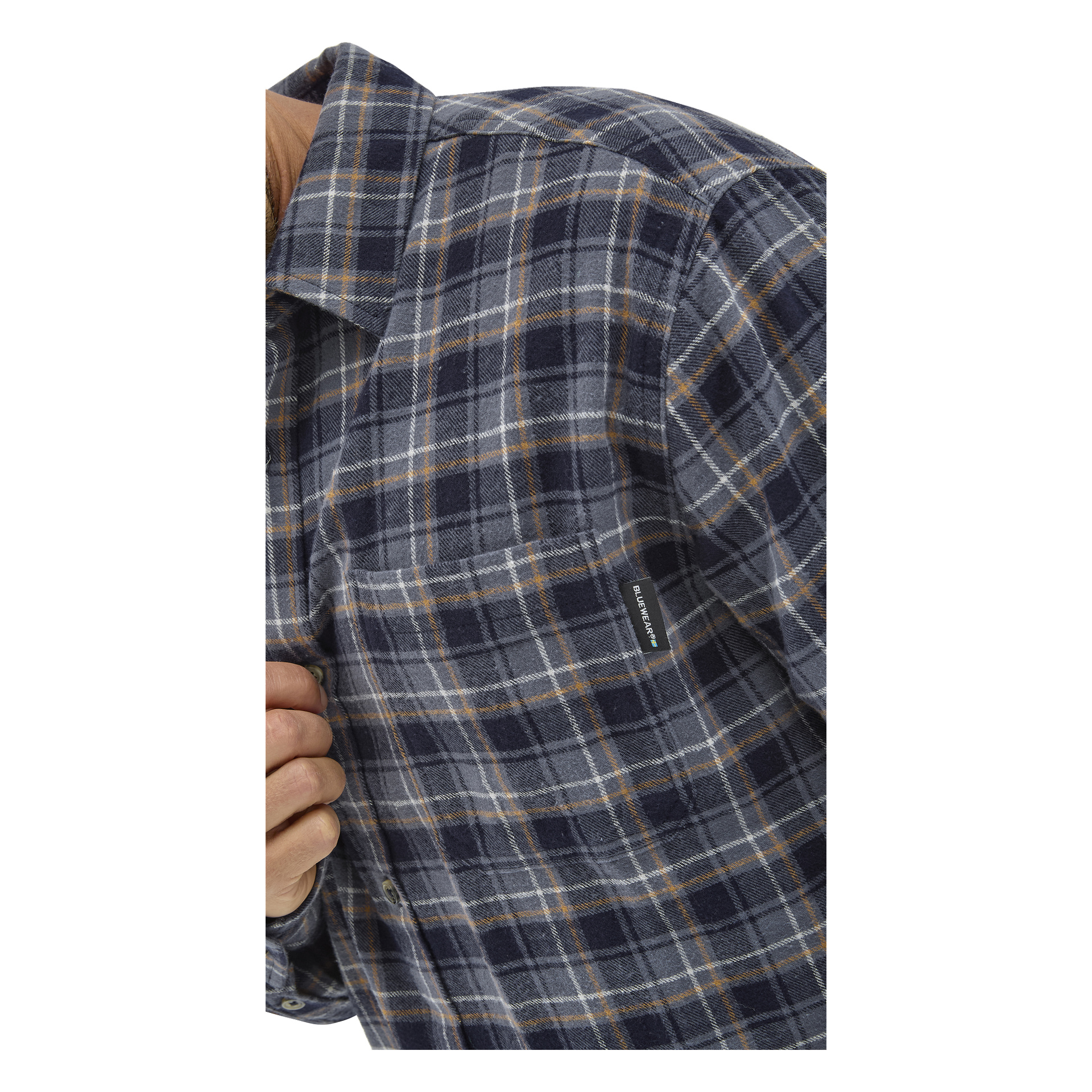 Närbild på en person som bär en blå, grå och gul rutig skjorta med knappar och en synlig bröstficka och en liten tagg nära fickan. Personen håller i en del av skjortan nära knapparna.