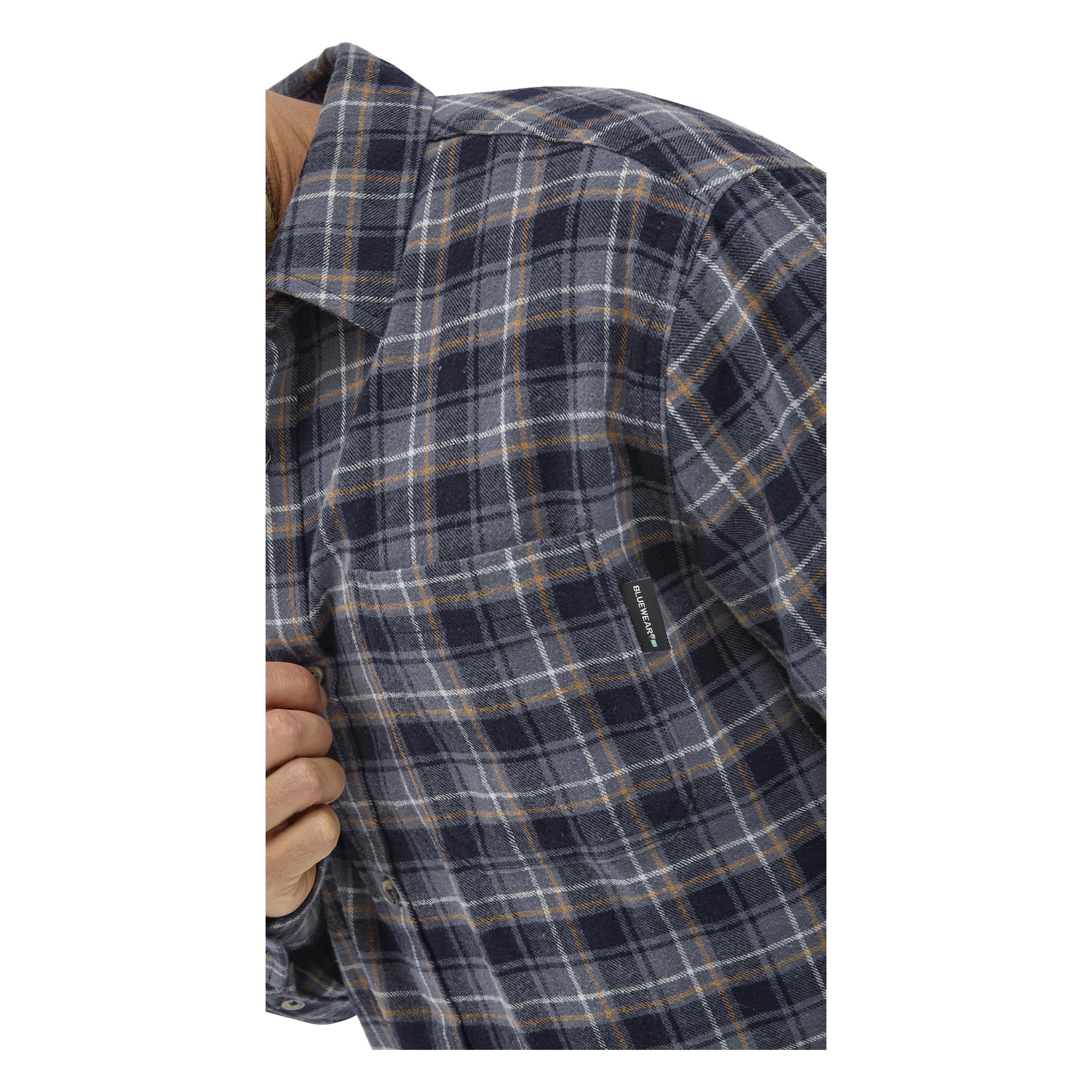 Närbild på en person klädd i en blå, grå och gul rutig flanellskjorta. En del av personens hand syns, som håller skjortan nära bröstfickan, som har en liten svartvit märkesetikett.