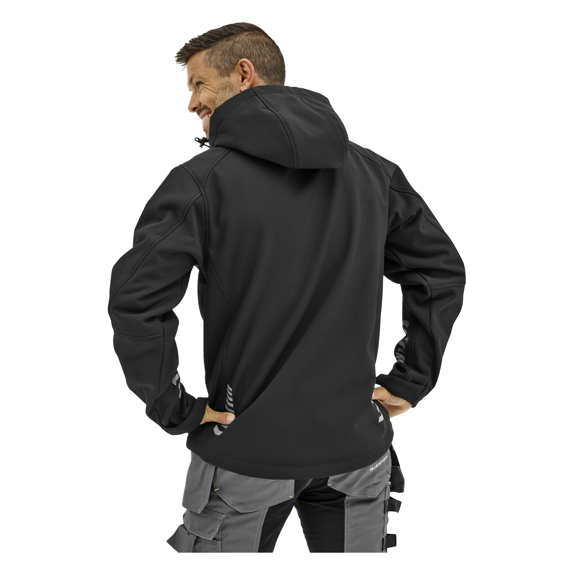 En man ler över axeln, klädd i en svart Bluewear Gustaf softshell-jacka och grå arbetsbyxor med fickor och öglor, händerna på höfterna.