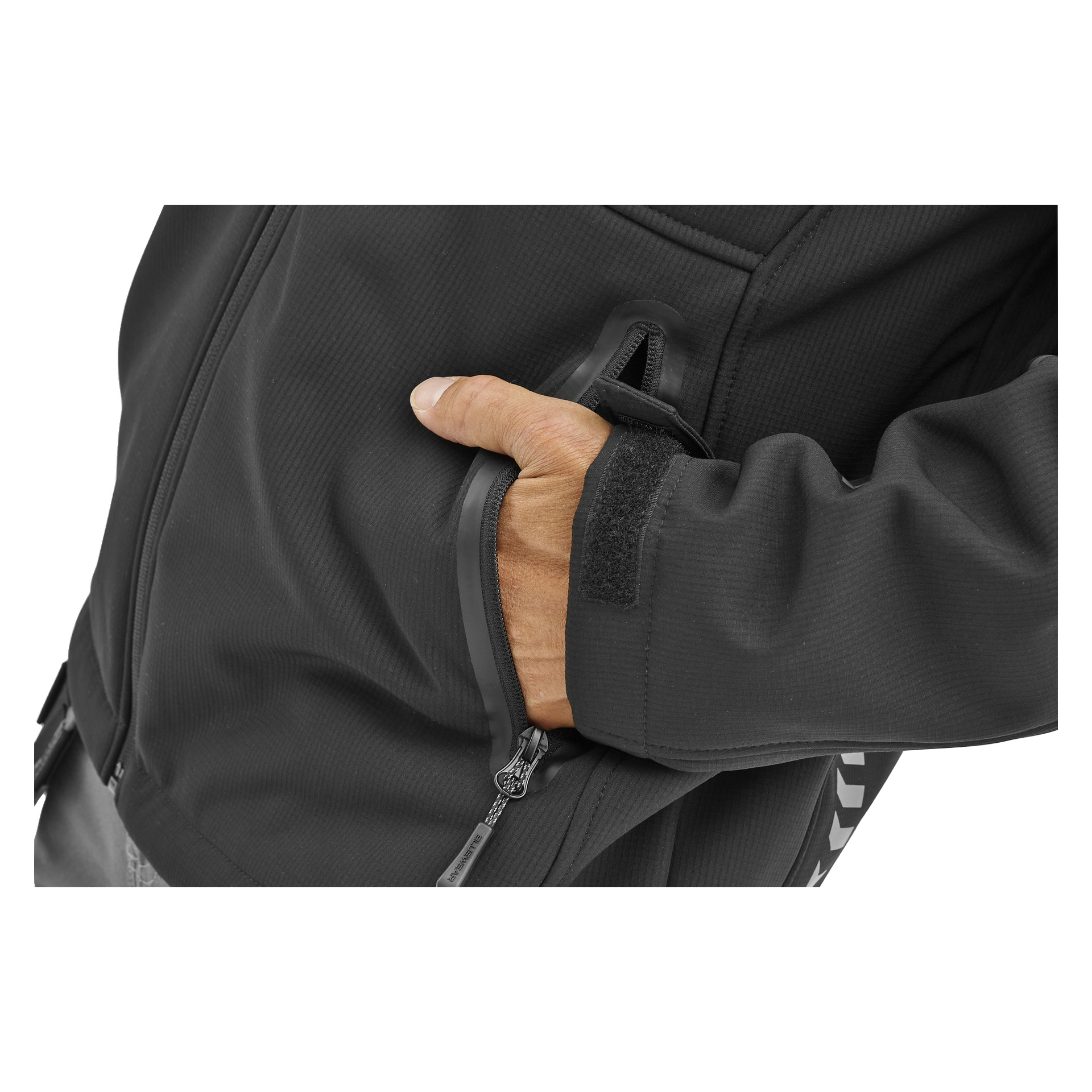 En man i en svart Bluewear Gustaf-softshelljacka drar ner dragkedjan på höger hand i fickan; ärmen har en hak-och-ögla-fästanordning.