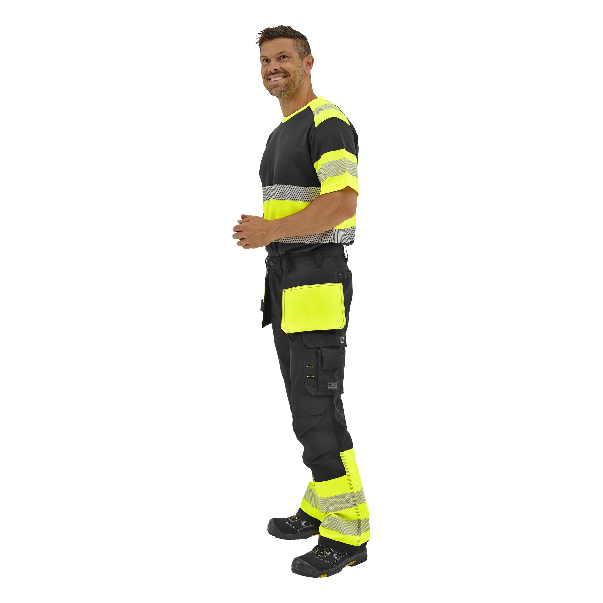 En man står på sidan i svart och neongult Bluewear High Visibility Trousers Rex och svarta arbetsskor, leende med sammanflätade händer.