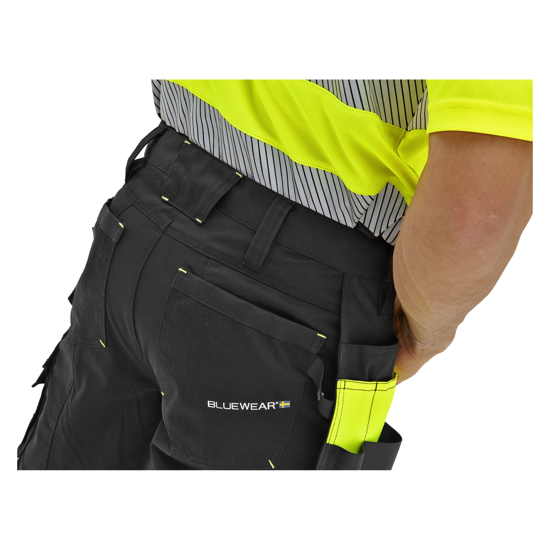 En person i Bluewear High Visibility Trousers Rex med gula detaljer och logga, iförd en gulgrå randig skjorta, handen i bakfickan.