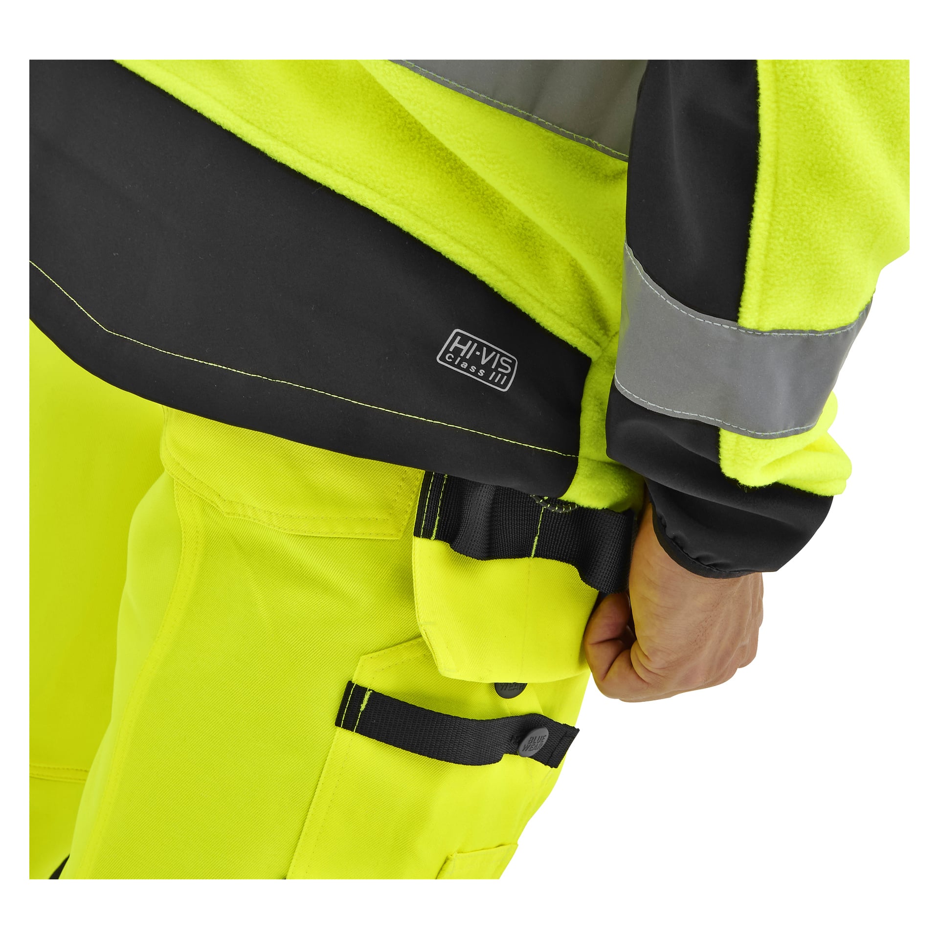 En person i en gul Bluewear Hi-Vis fleecejacka med svarta, reflekterande detaljer och en HI-VIS klass III-etikett justerar sina arbetsbyxor.