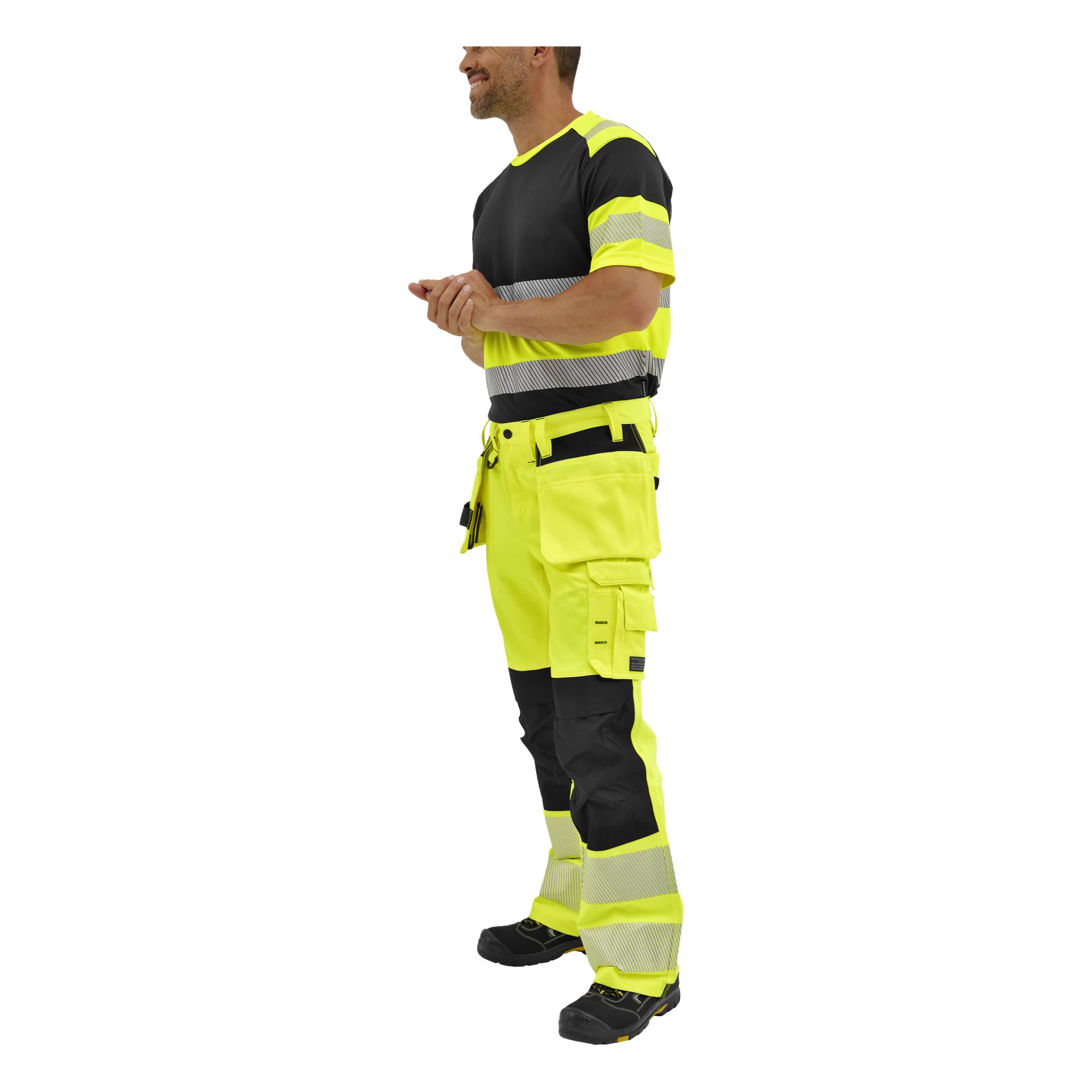 En man står vänd åt vänster, klädd i Bluewear Rex hög-vis-byxor med reflekterande ränder, en svart-gul hög-vis-skjorta och arbetsskor.
