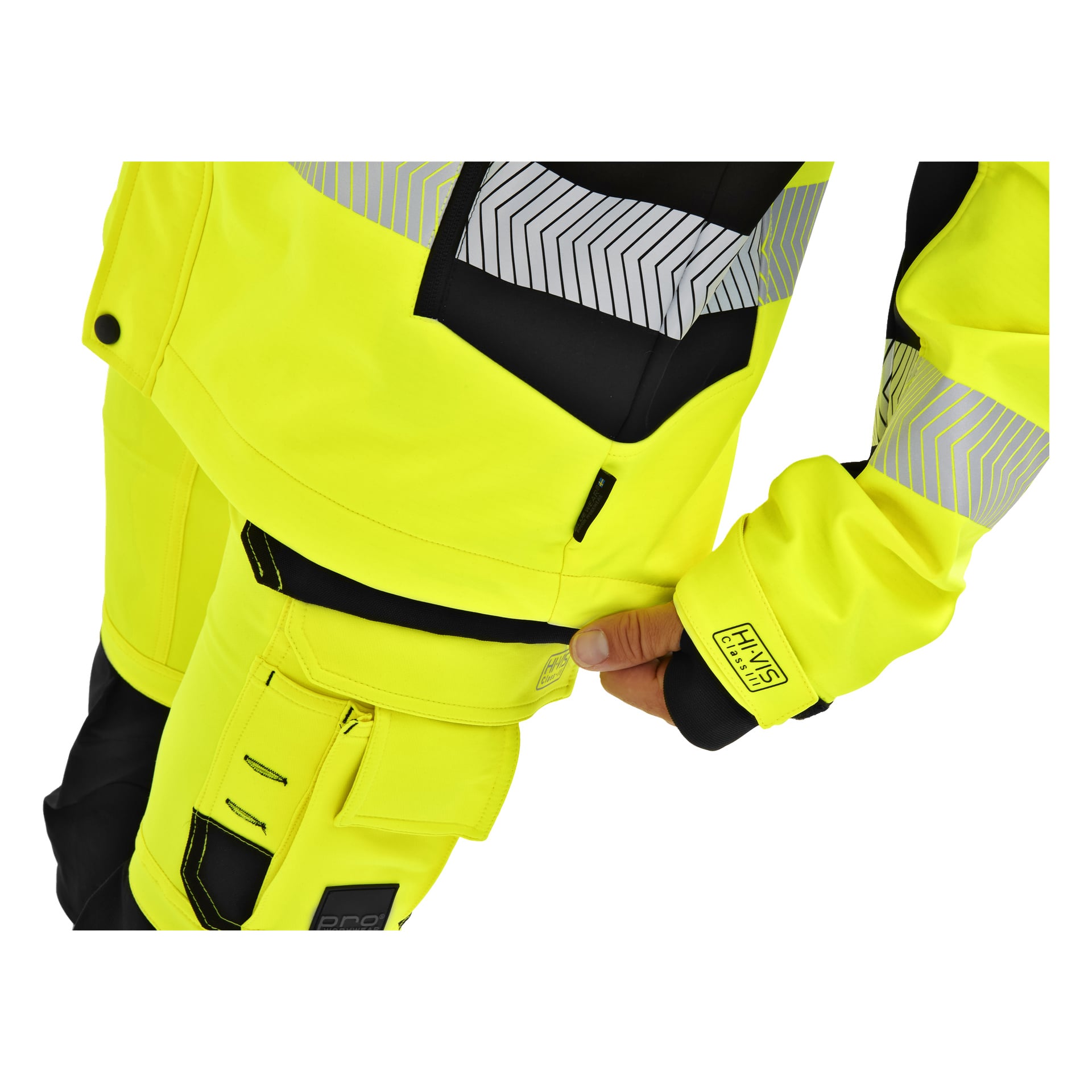 Man justerar muddringen på en gul Bluewear Hi-Vis softshelljacka med svarta och reflekterande detaljer, som visar fickor och dragkedjor.