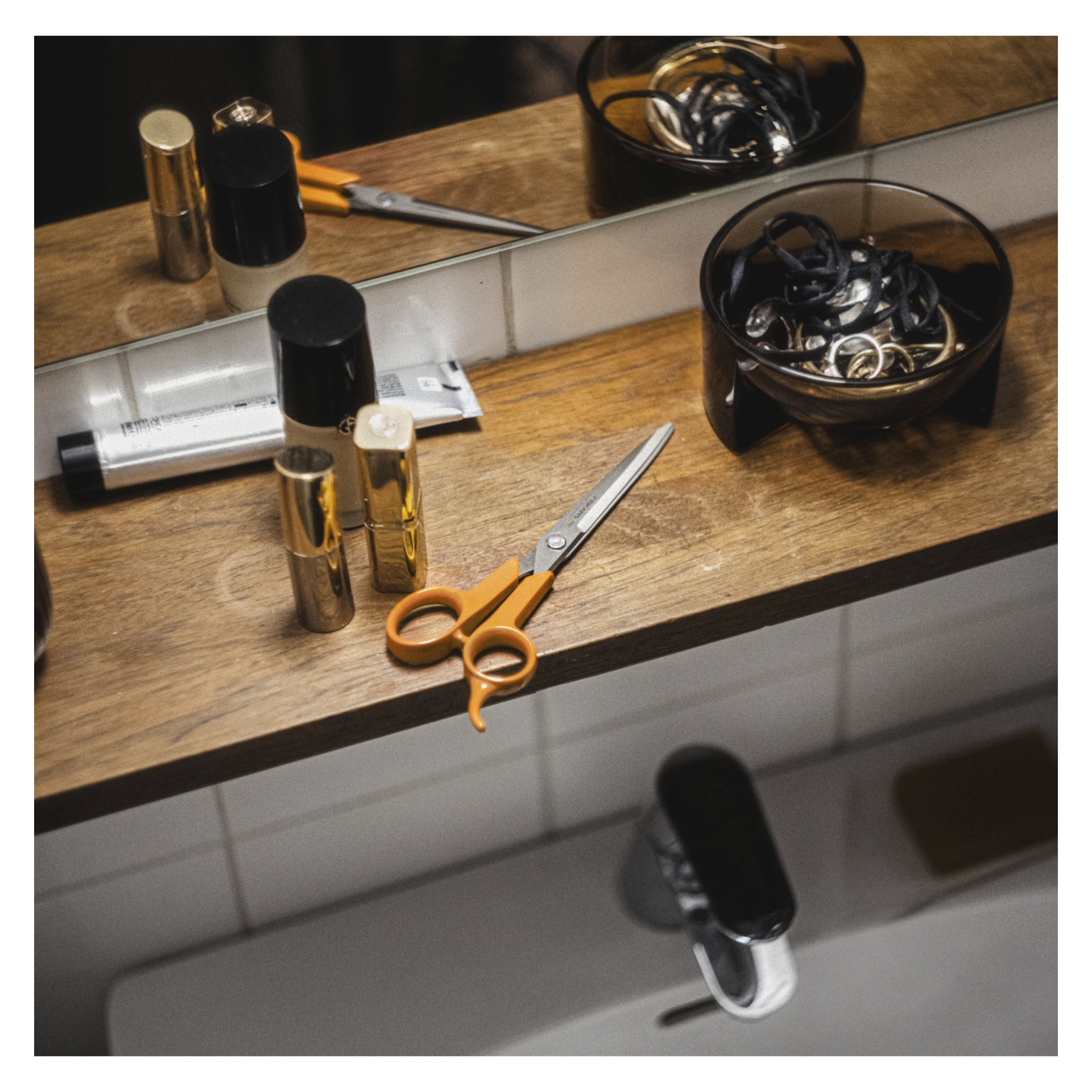 På en badrumshylla i trä ovanför ett handfat finns en sax med orange handtag, hårsnoddar i en mörk skål samt olika flaskor och tuber med hygienprodukter. En spegel reflekterar föremålen på hyllan.