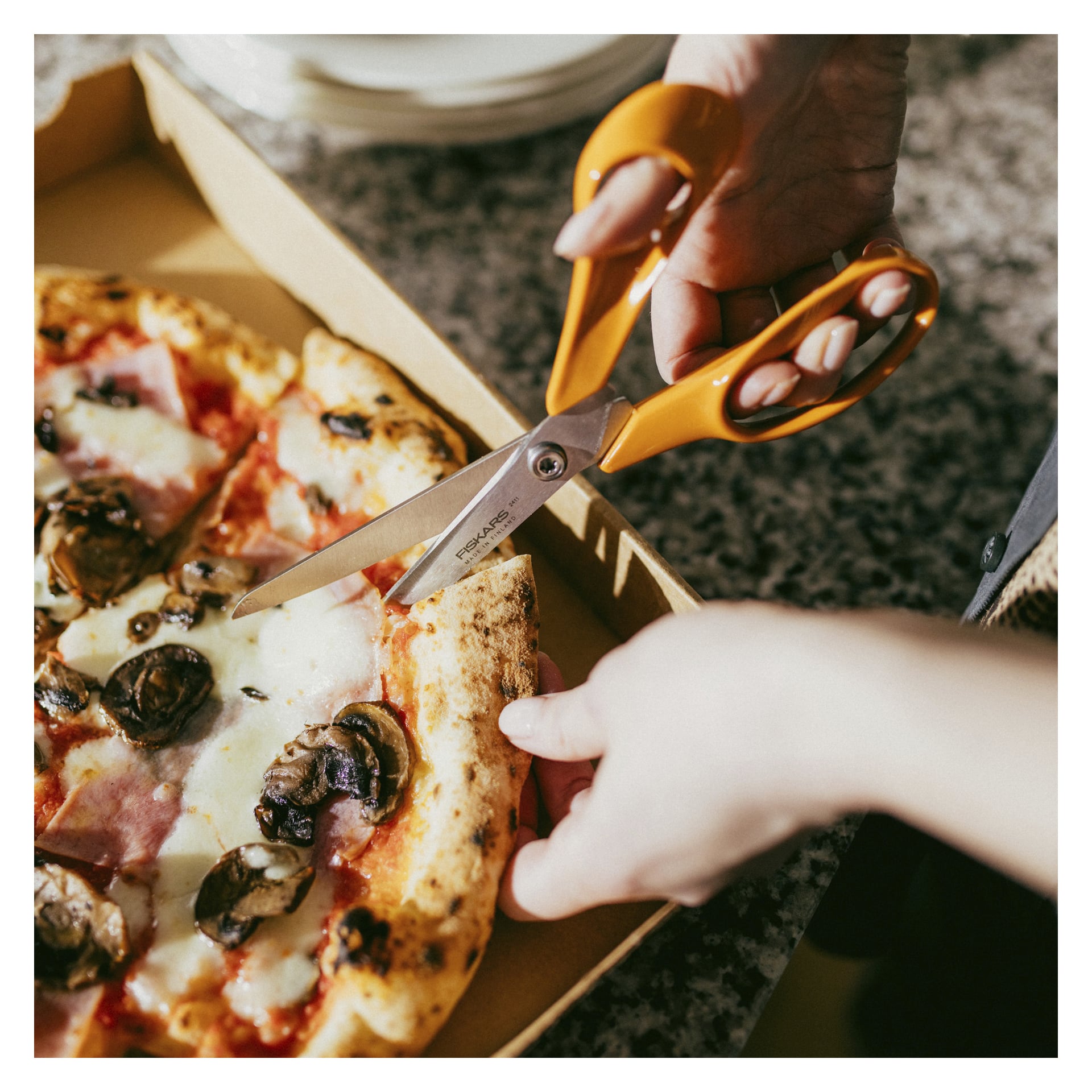 Eine Person schneidet mit einer orangefarbenen Schere ein Stück von einer Pizza mit Käse, Pilzen und Schinken ab, die in einem Karton auf einer gesprenkelten Arbeitsfläche liegt.