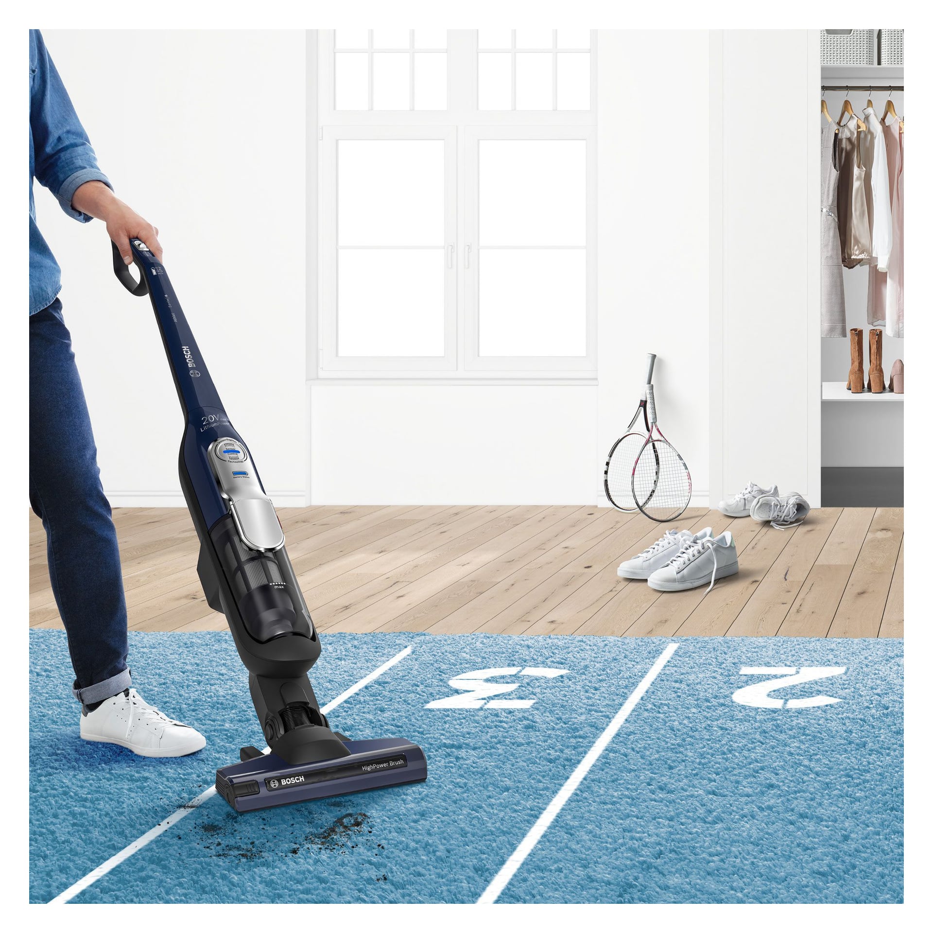 En person dammsuger en blå matta med vita spårlinjer med en dammsugare från Bosch. I närheten finns ett tennisracket, vita skor och hängande kläder i ett ljust, modernt rum.