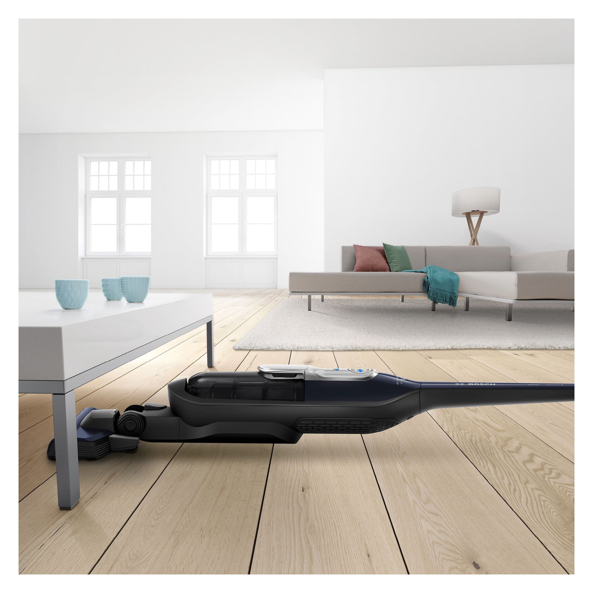 En sladdlös dammsugare rengör ljusa trägolv under ett vitt soffbord i ett ljust, modernt vardagsrum med soffa, kuddar, lampa och dekorativa koppar.