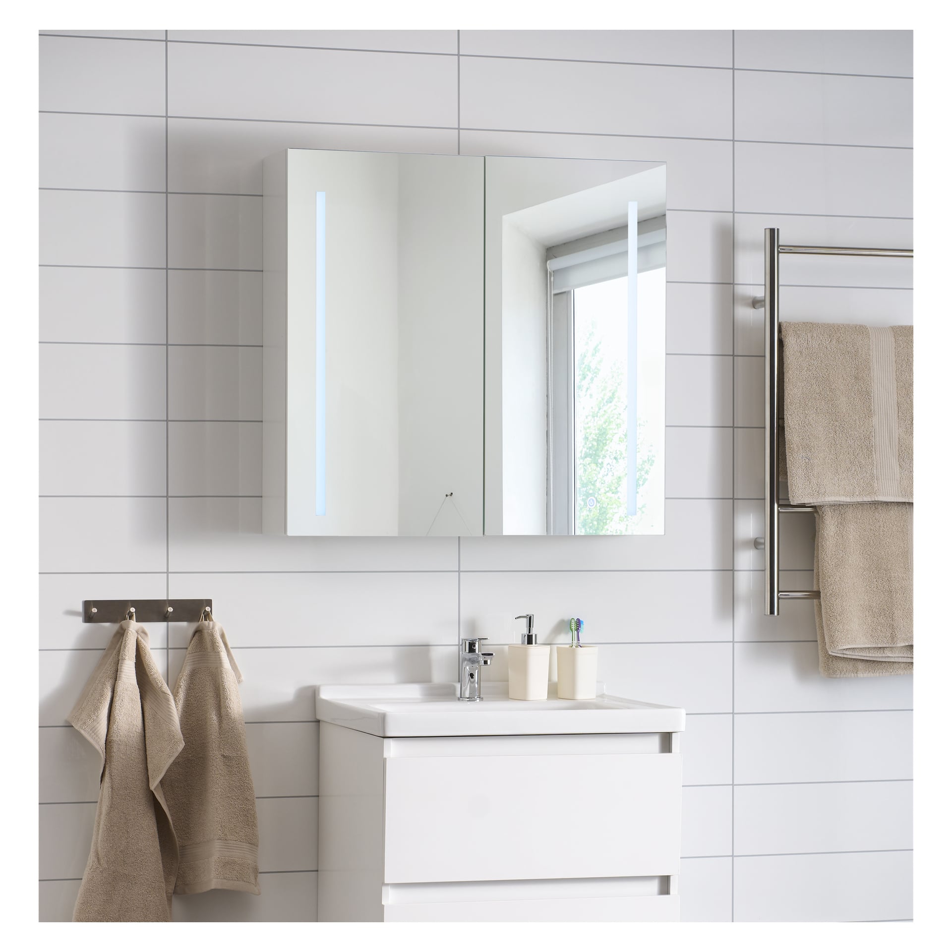 Modernt badrum med vitt kakel, ett spegelskåp över ett vitt handfat, handdukshängare i krom med beige handdukar och ett fönster som visar grönska.