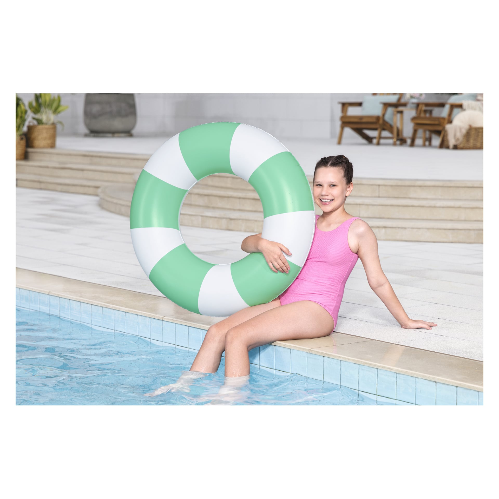 En flicka i rosa baddräkt sitter på kanten av en pool med fötterna i vattnet och håller i en grön och vit uppblåsbar simring. I bakgrunden syns en trappa och stolar på uteplatsen.