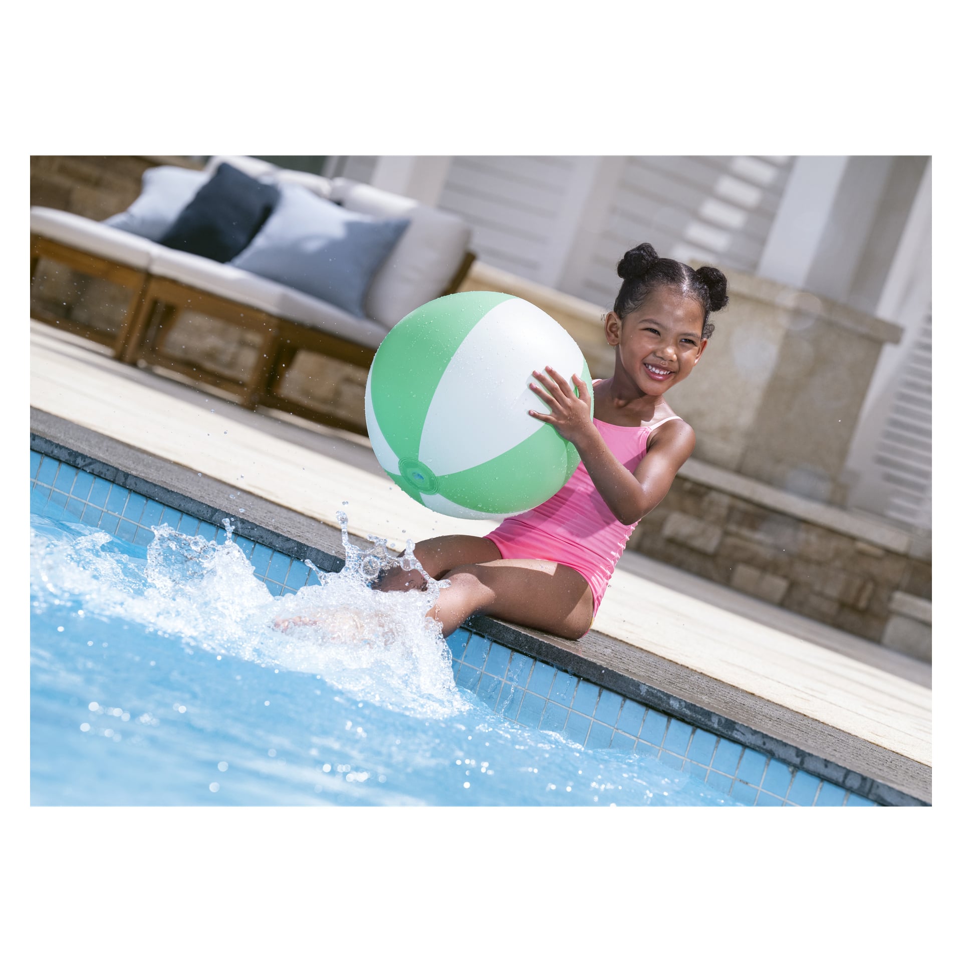En ung flicka i rosa baddräkt sitter vid kanten av en pool, håller i en grön och vit badboll, ler och plaskar med fötterna i vattnet. I bakgrunden finns en uteservering och en solig miljö.
