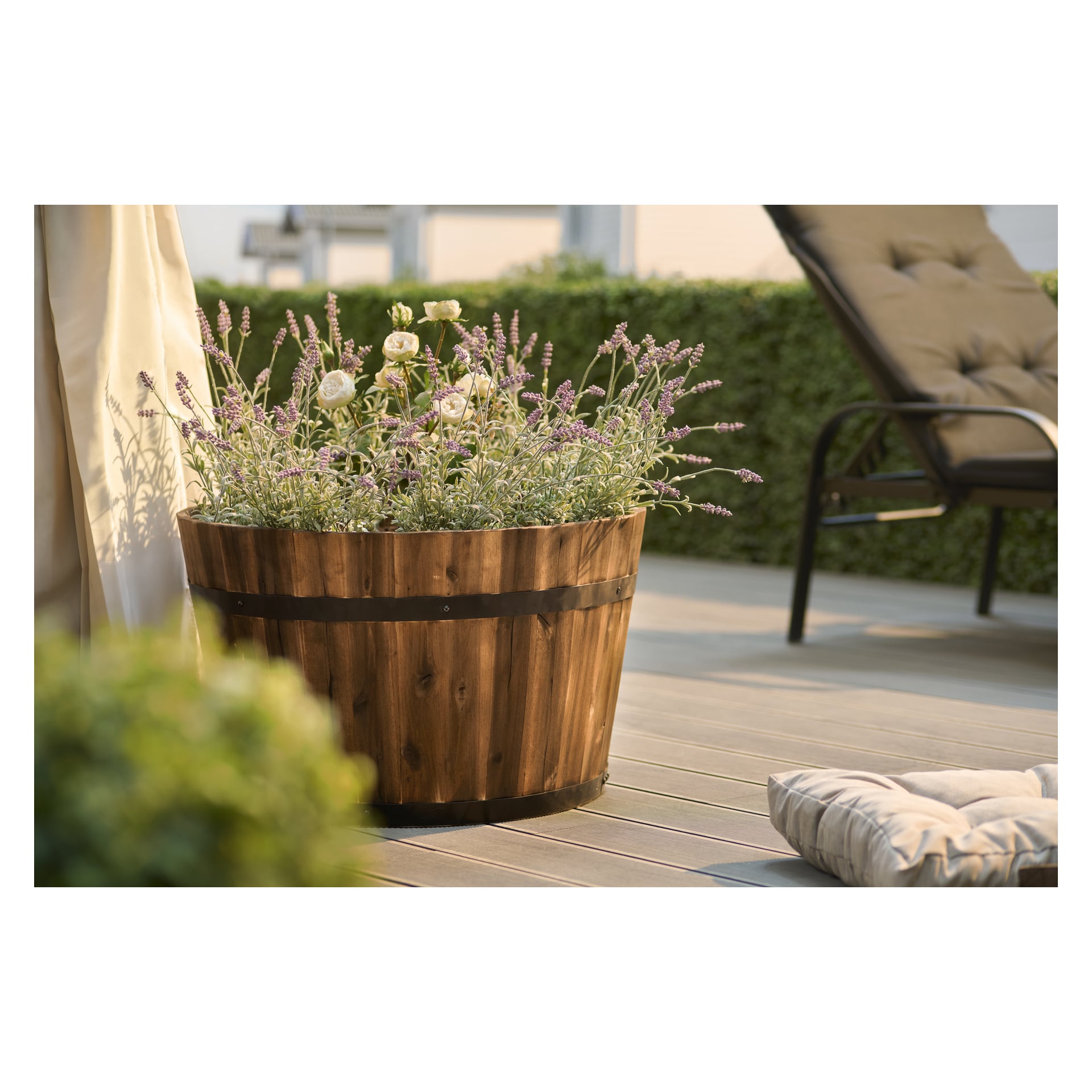 En träkruka med lavendel och vita blommor står på en ljus terrass nära en solbränd fåtölj med kuddar. I bakgrunden skymtar gröna häckar i det starka solljuset.