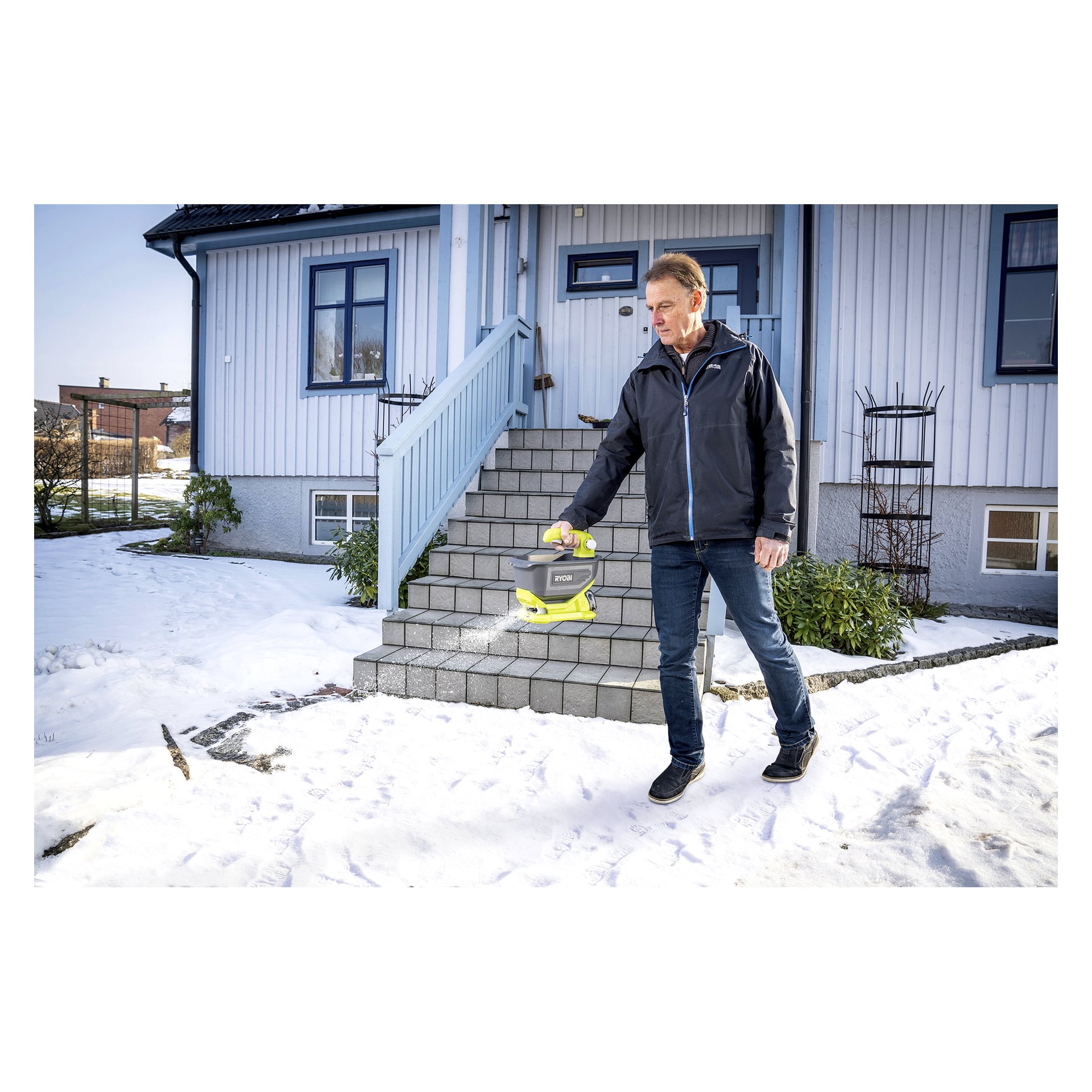 En man använder en handhållen maskin för att sprida material på snöiga trappsteg utanför ett hus, sannolikt för att smälta is eller ge grepp. Huset och gården är täckta av snö och växter syns nära trappan.