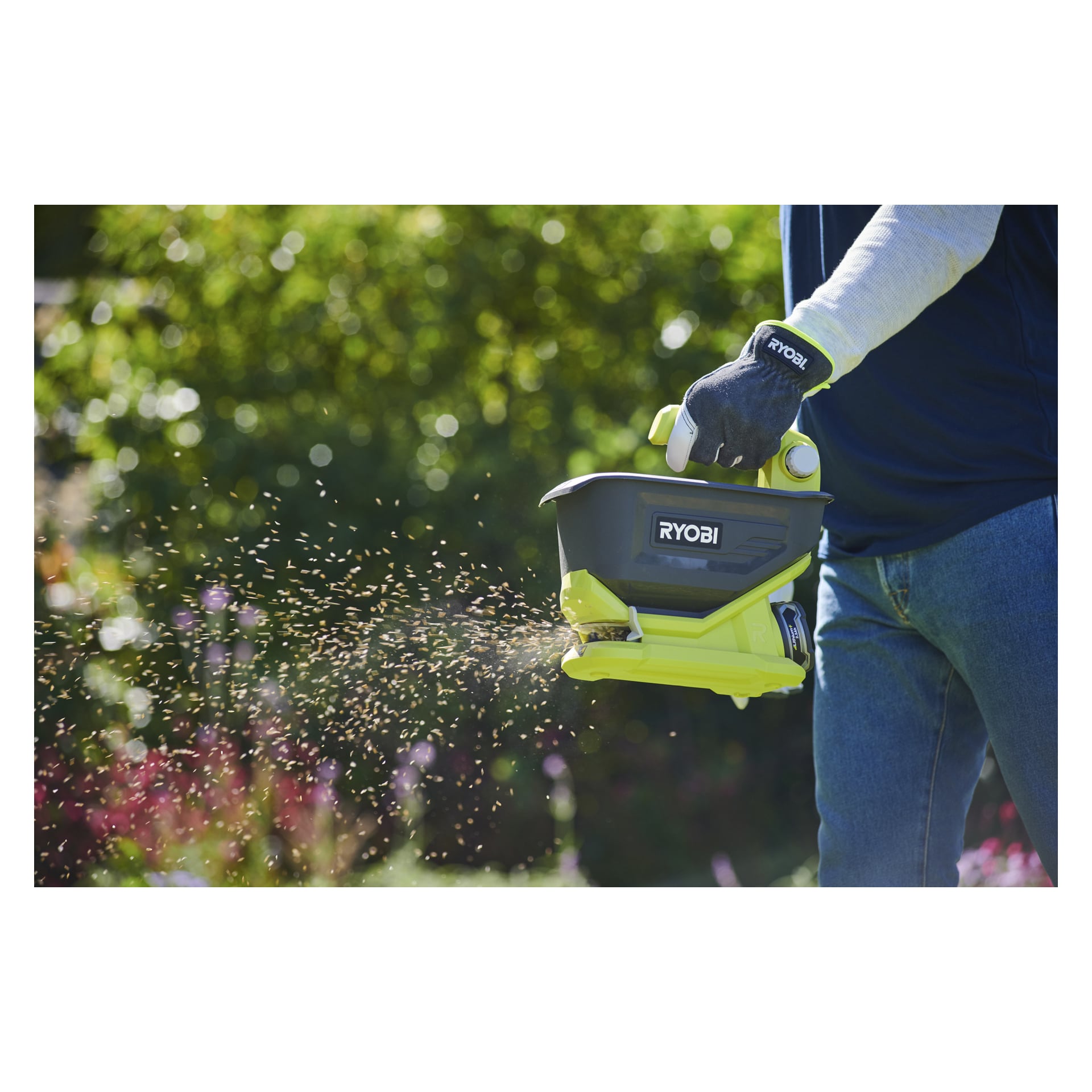 En person med handskar använder en handhållen fröspridare av märket Ryobi i en trädgård och sprider små frön eller gödningsmedel på marken. Frodig grönska och blommande växter syns i bakgrunden.