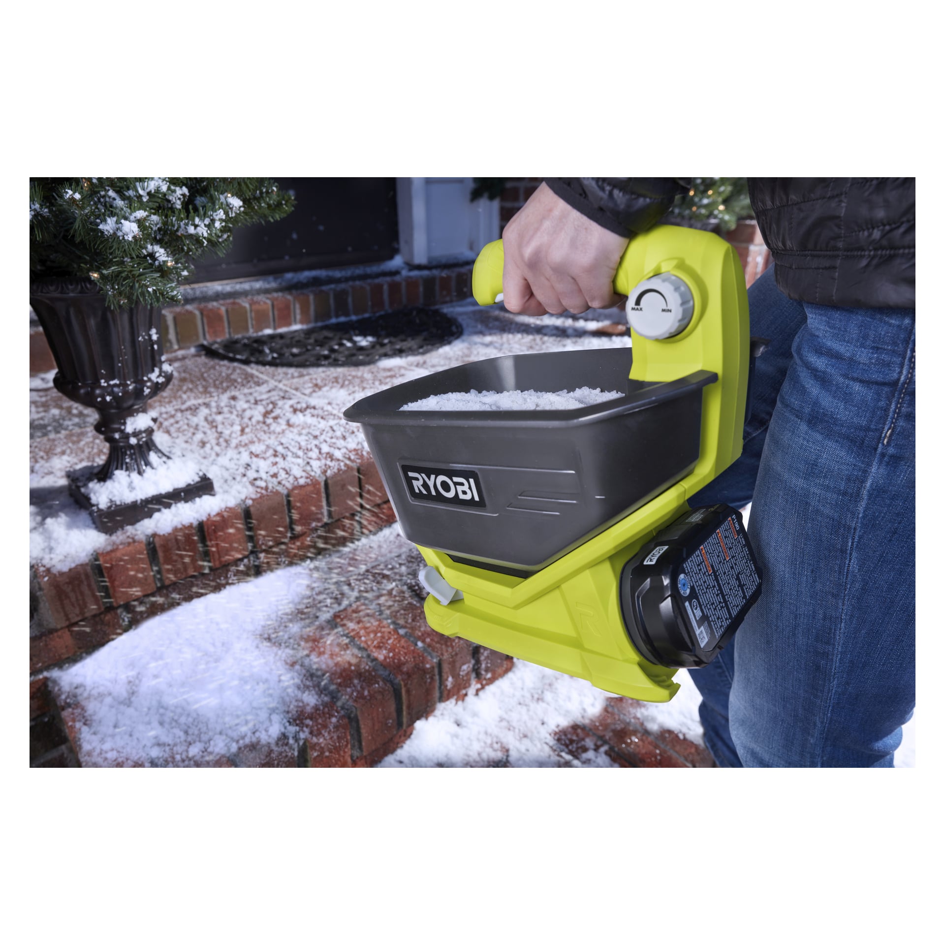 En person håller i en gul Ryobi handhållen spridare fylld med vitt granulat och sprider det på snötäckta tegeltrappor framför ett hus.