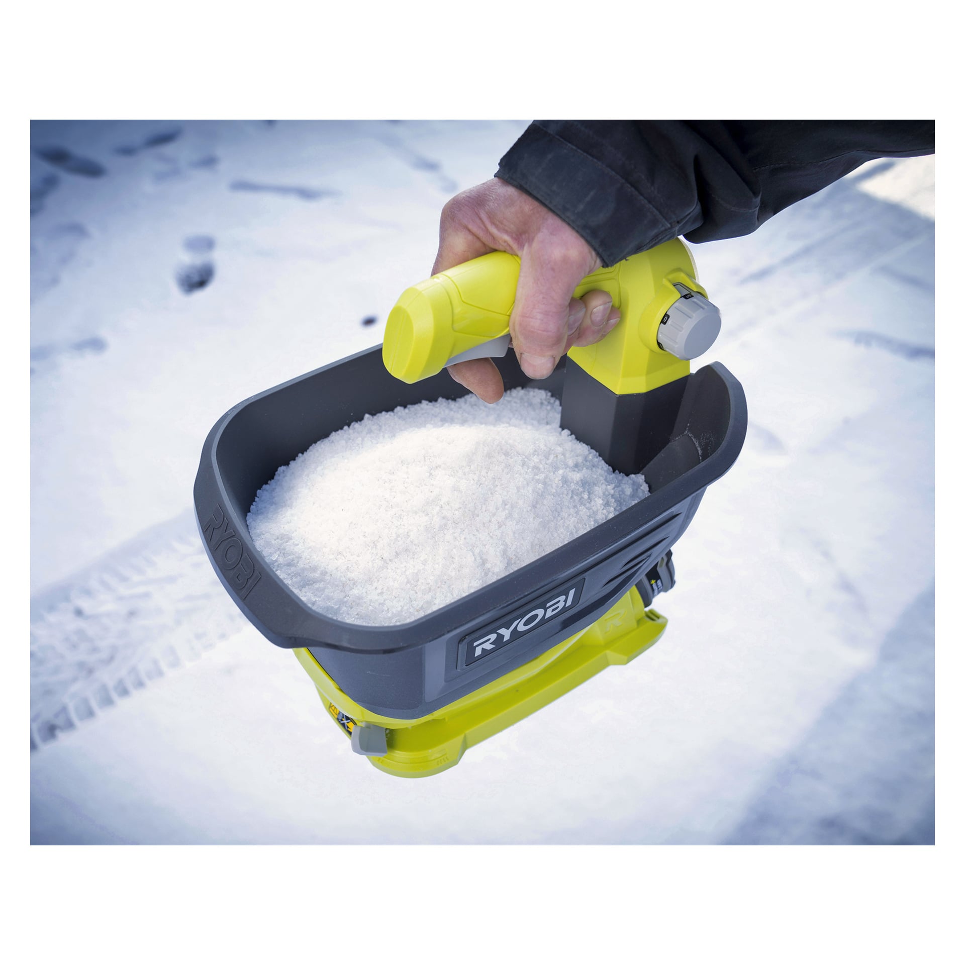 En hand håller en gul och svart Ryobi handhållen spridare fylld med vita saltkristaller, med snötäckt mark synlig i bakgrunden.