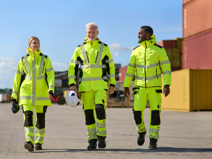 Tre personer iført gule arbeidsklær med høy synlighet går utendørs på et asfaltert område i nærheten av containere og stablet last. Teksten Nyheter vises i en gul boks i øvre venstre hjørne.
