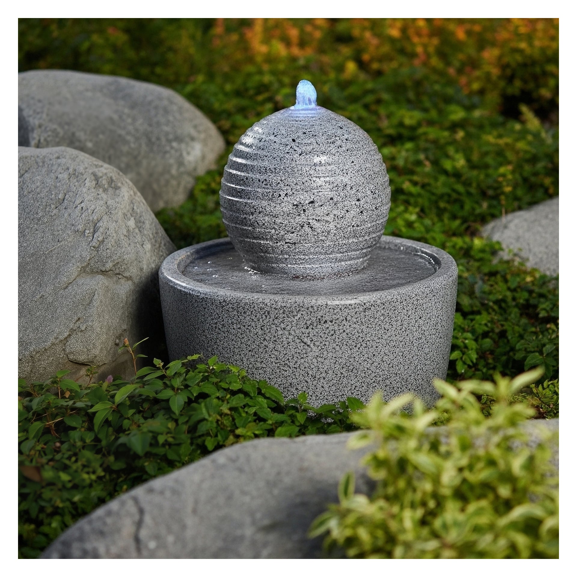 En liten, rund Hard Head Fountain med LED-lampor har en ribbad sfär på toppen och står bland gröna växter och stora grå stenar. Vatten rinner från toppen av sfären och skapar krusningar i bassängen nedanför.
