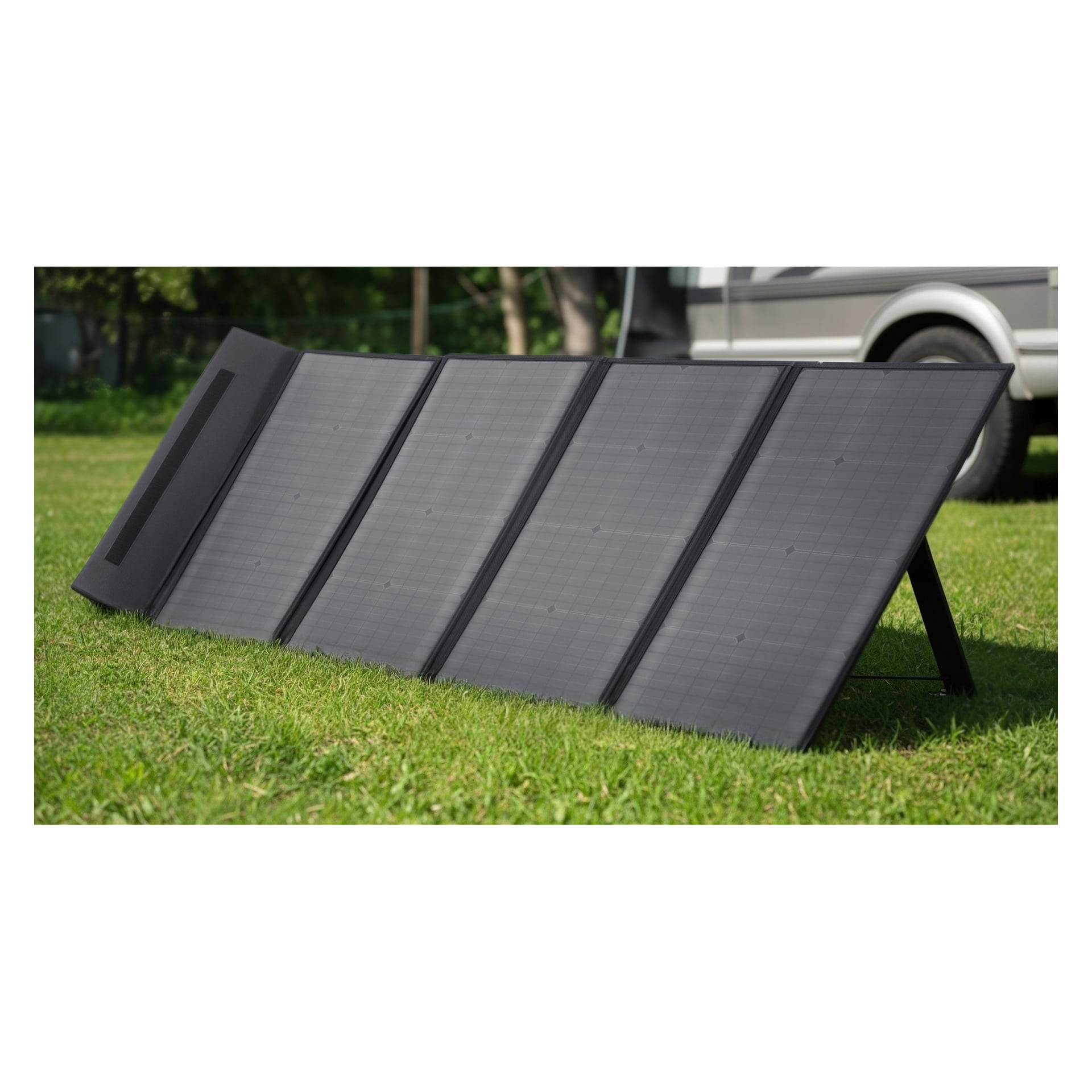 Ett hopfällbart svart Hamron Solarcell-paket står på grönt gräs utanför, med träd och en del av ett närliggande parkerat fordon bakom.