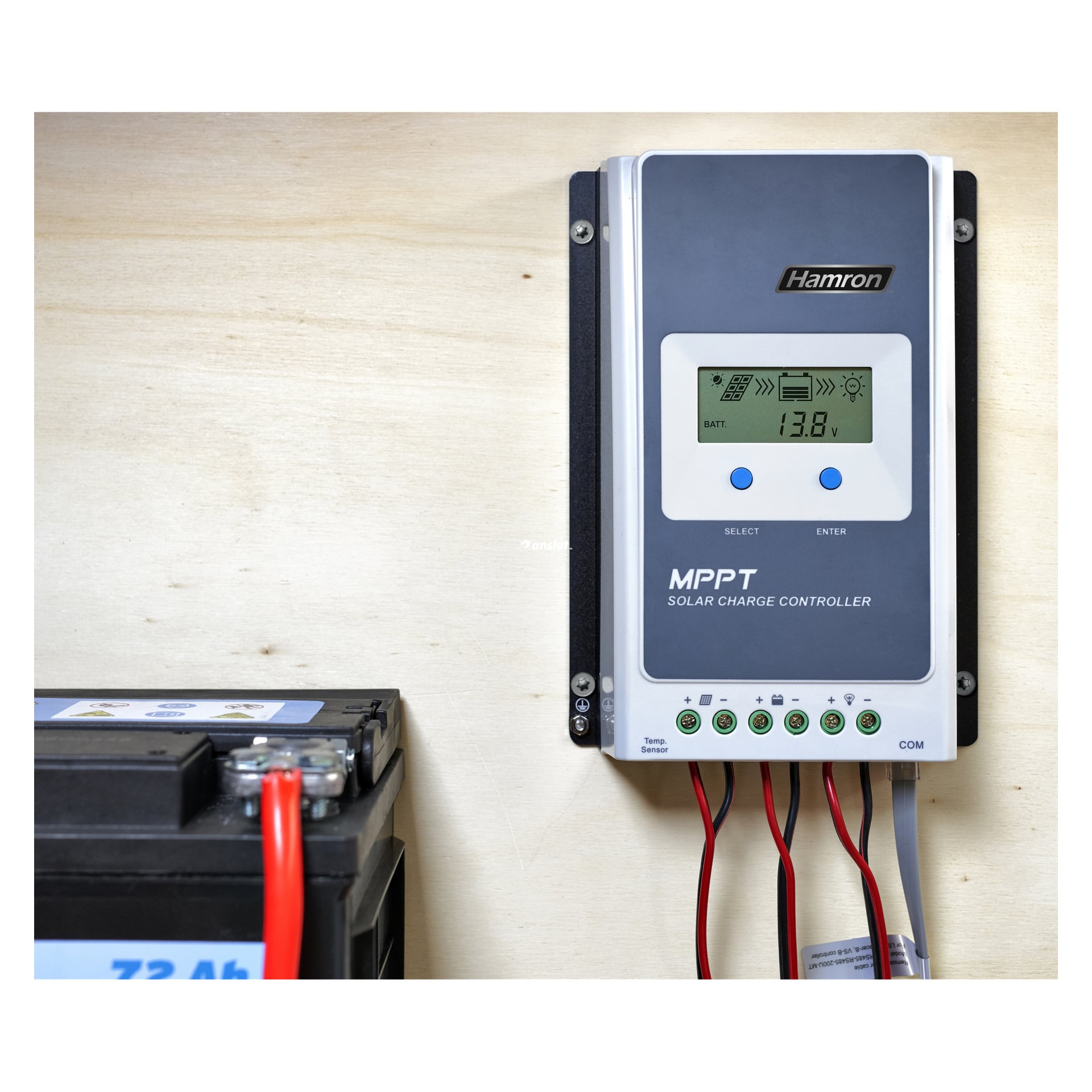 En Hamron Charging-regulator är fäst vid en ljus trävägg och visar information på skärmen, med kablar nedanför och ett batteri i närheten.