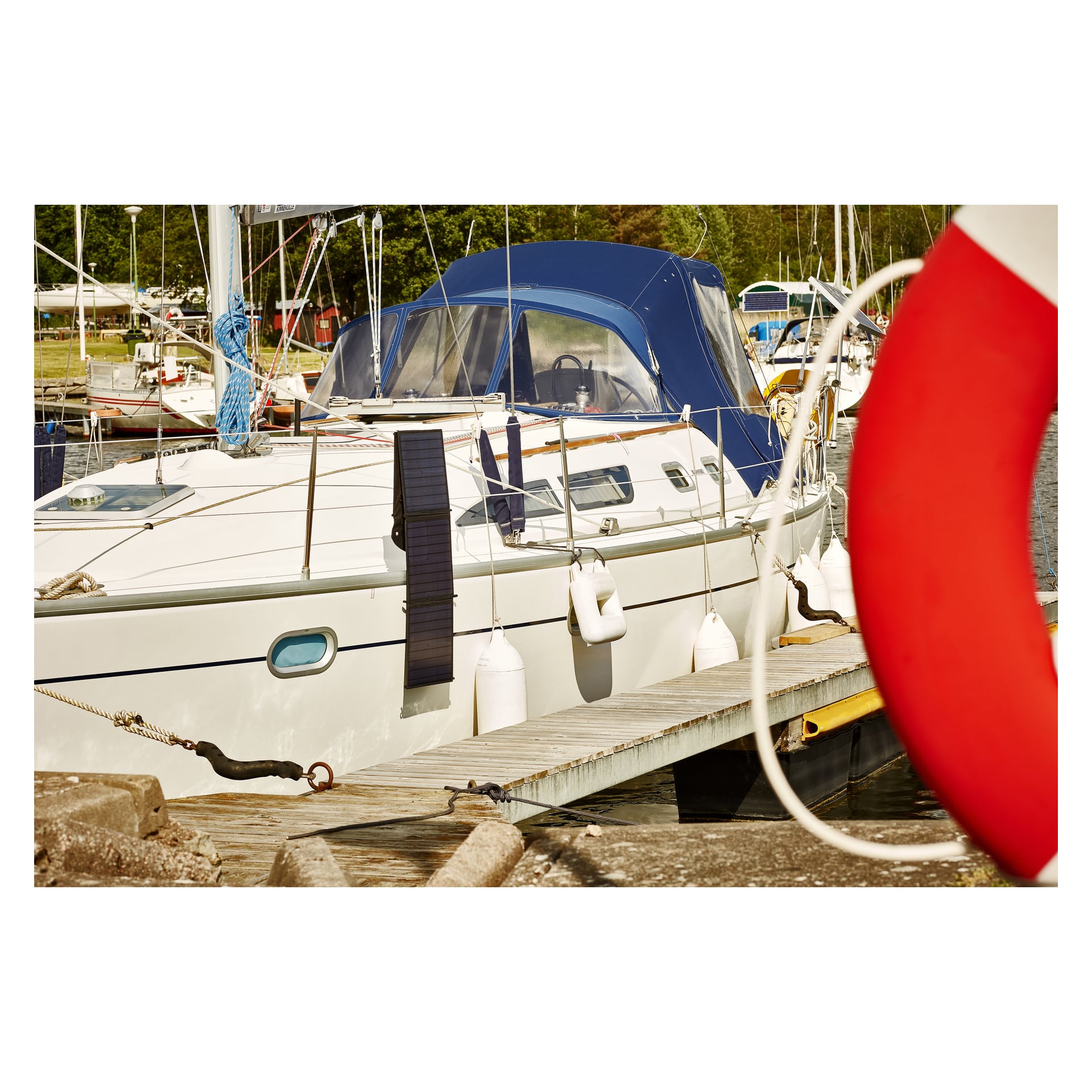 En Hamron-solpanel och regulator finns på en vit segelbåt vid en marina, med fendrar och en rödvit livboj synliga i närheten.