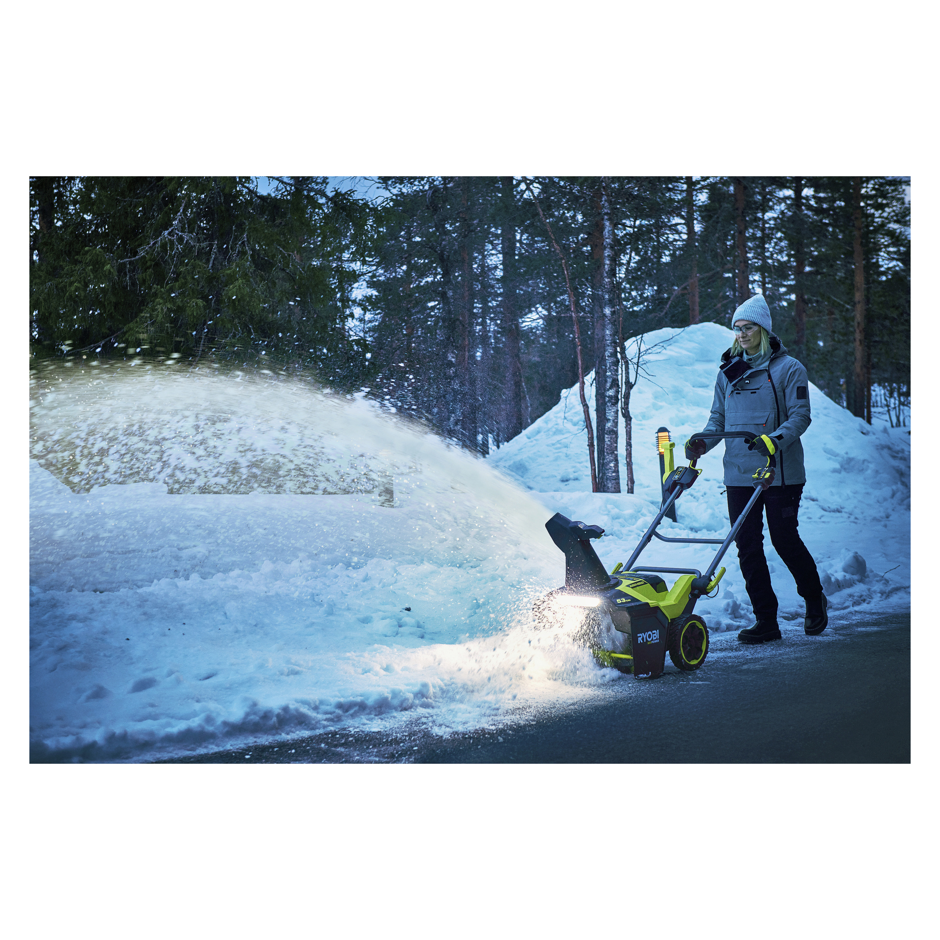 En person i vinterkläder kör en Ryobi Max Power-batteri snöslunga och rensar en snötäckt uppfart omgiven av träd och snödrivor.