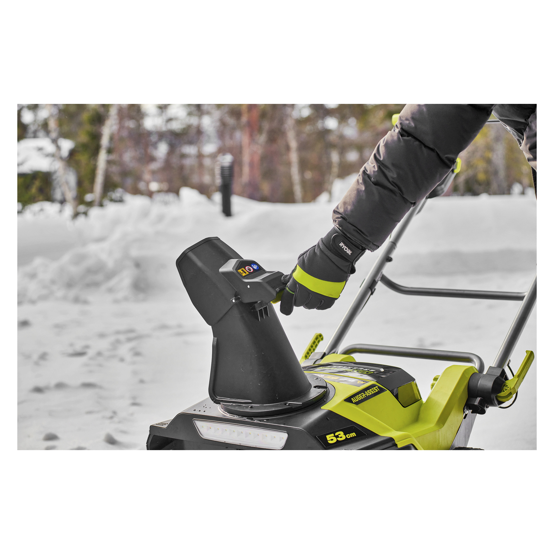 Handskbeklädda händer justerar fallskärmsspaken på en Ryobi Max Power-batteri utanför, omgiven av snö, träd och snödrivor.