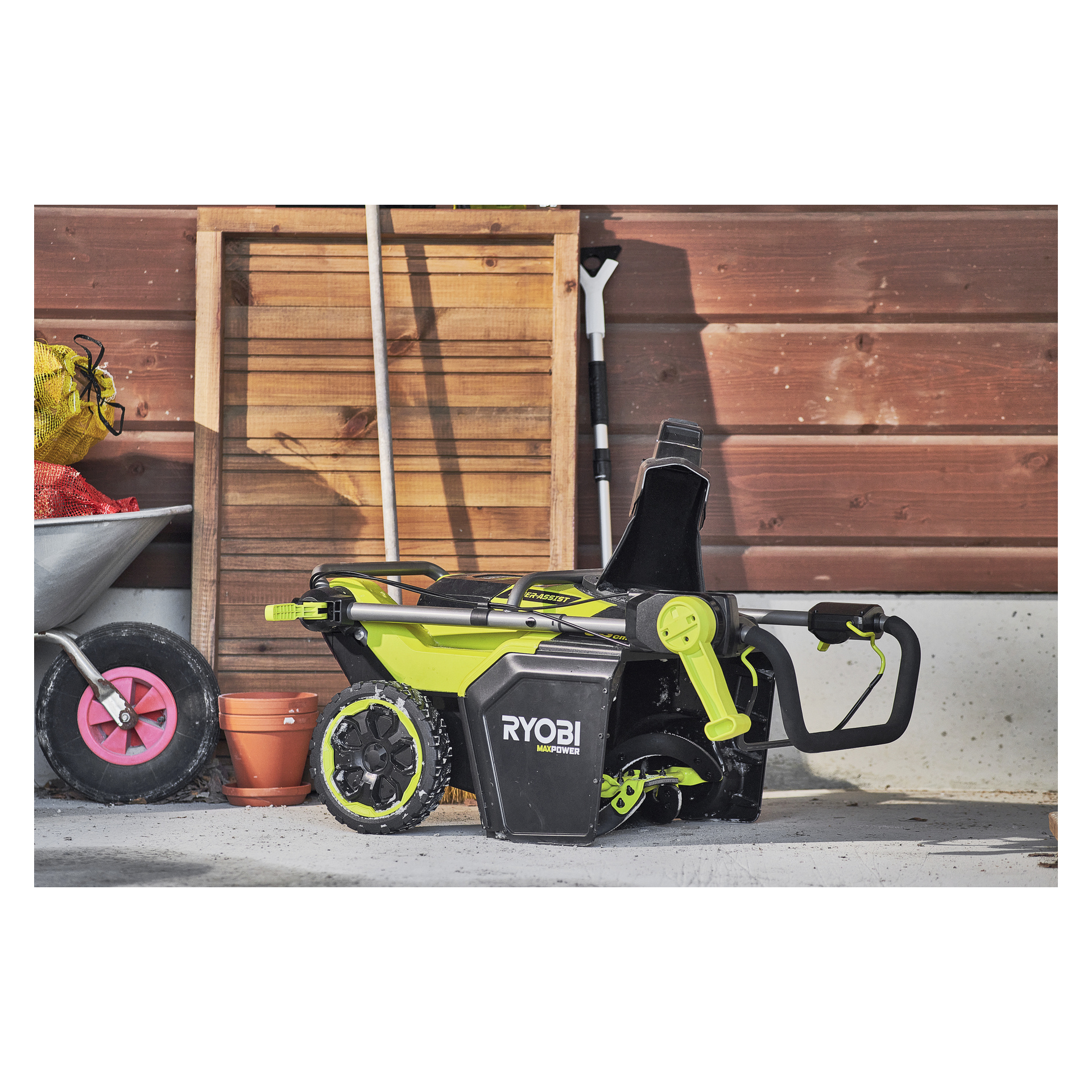 En Ryobi Max Power snöslunga RY36STX53B-160 ligger på sidan i ett garage vid en träpanel, skottkärra, blomkruka och nätpåse.